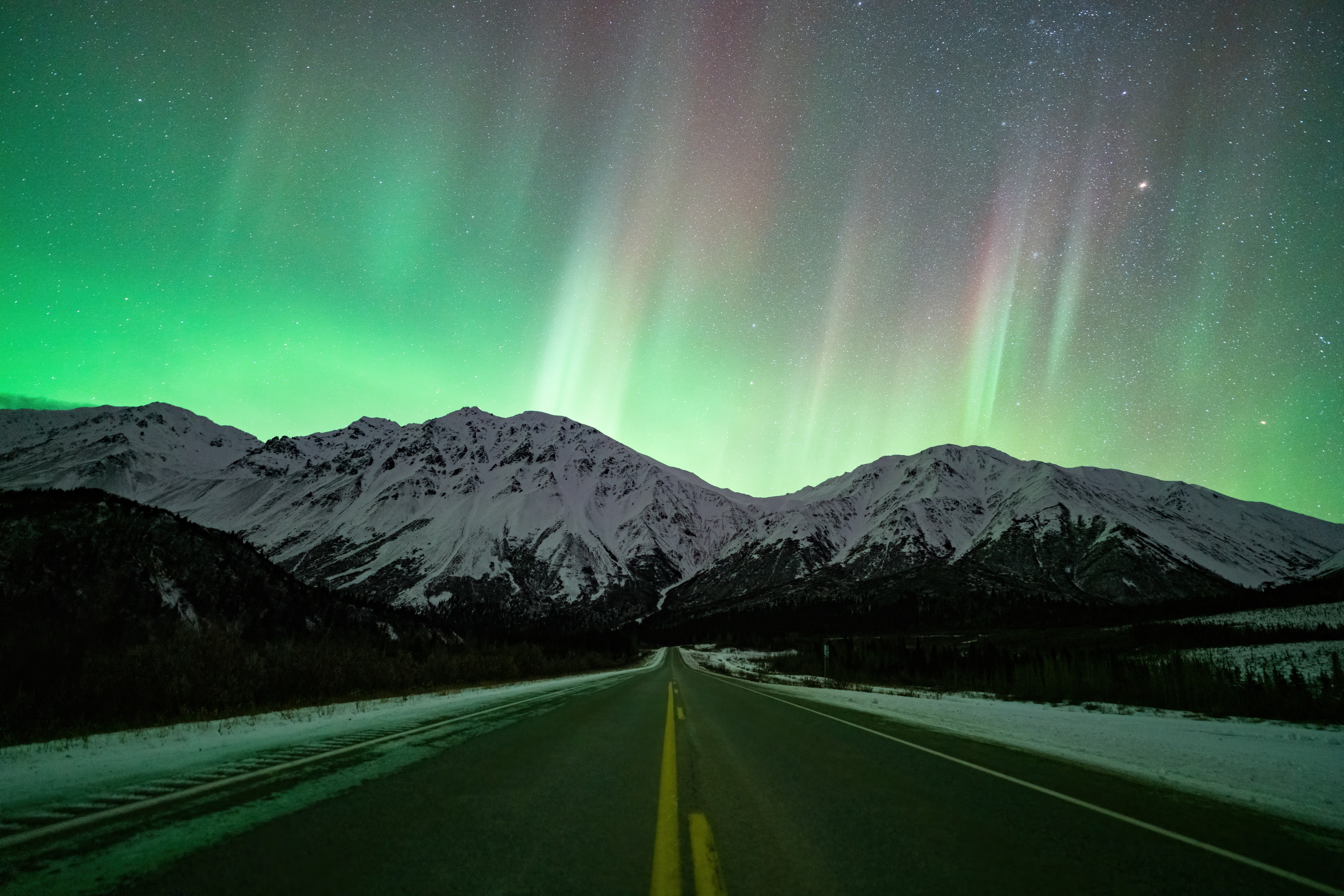 Nighttime landscape with snow-covered mountains and green aurora in the sky, viewed from a straight road.