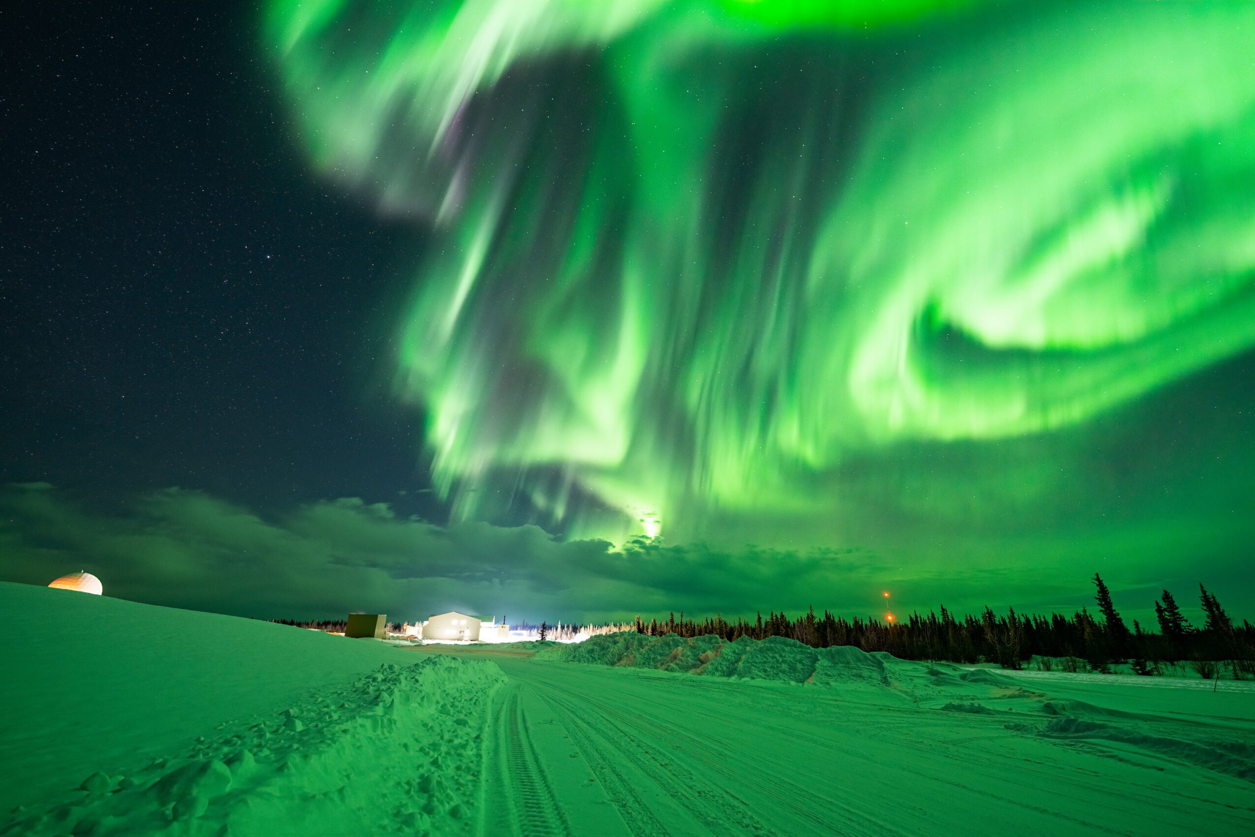 Northern lights display green aurora in night sky over snow-covered landscape with trees and distant lights.