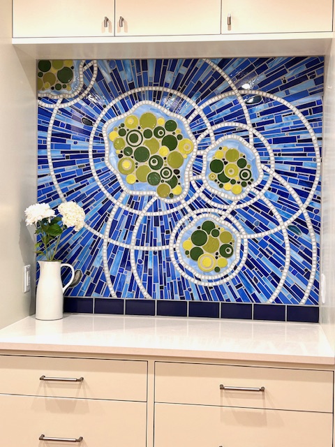 Colorful mosaic with blue, green, and yellow circular patterns on a wall, with a white vase of white flowers on a countertop.