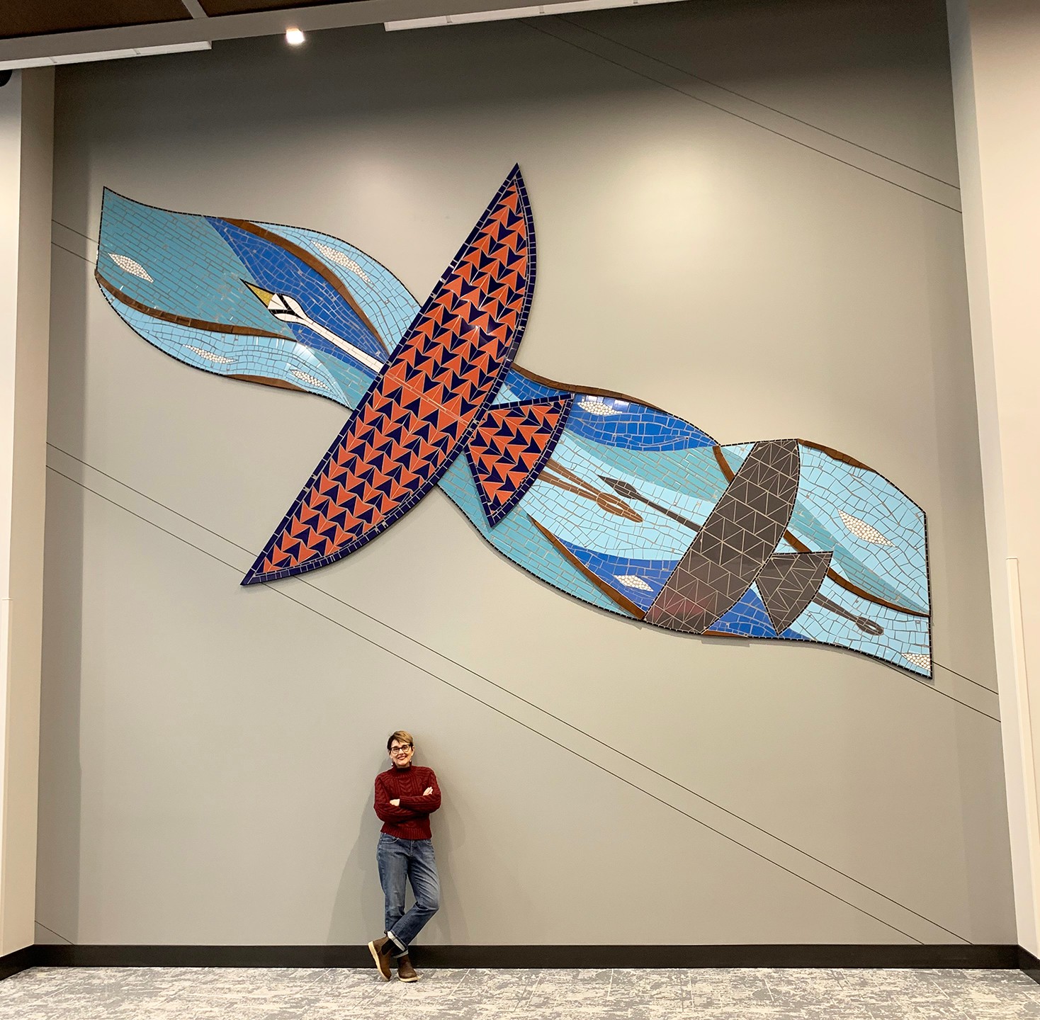 Person standing in front of a large, colorful abstract wall art resembling a bird with blue, orange, and gray patterns.