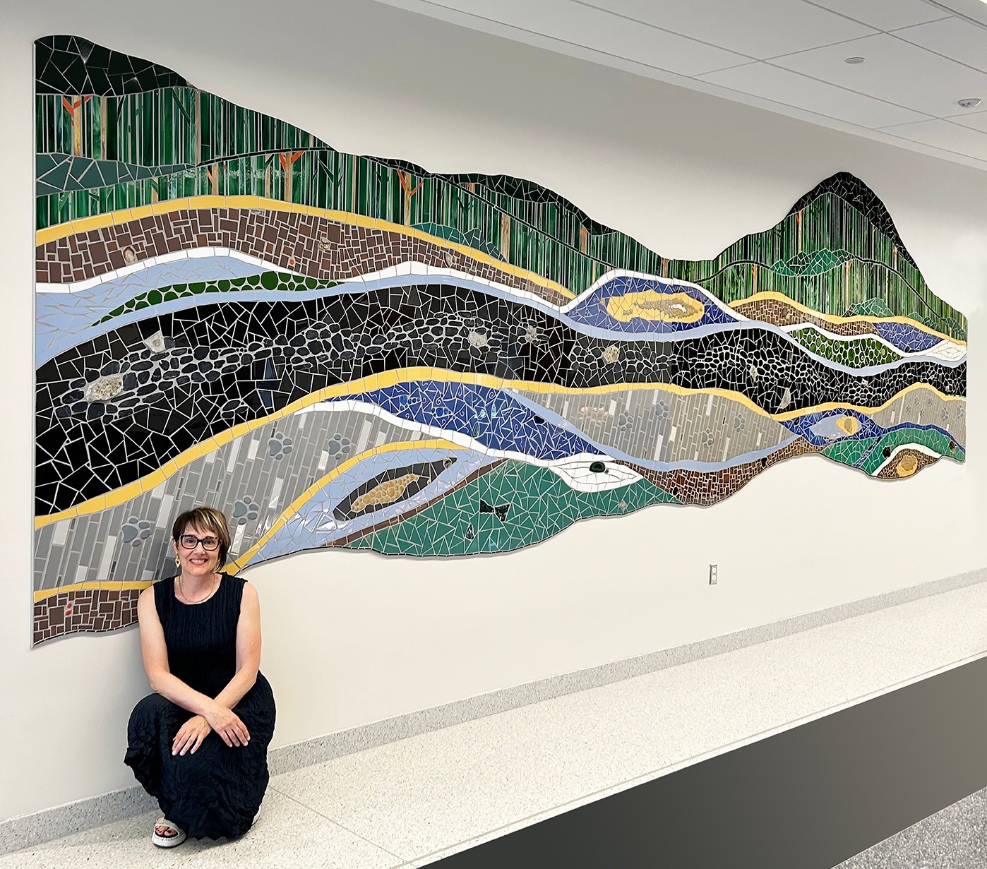 Person sitting on floor in front of a large colorful mosaic wall art depicting abstract landscape patterns.