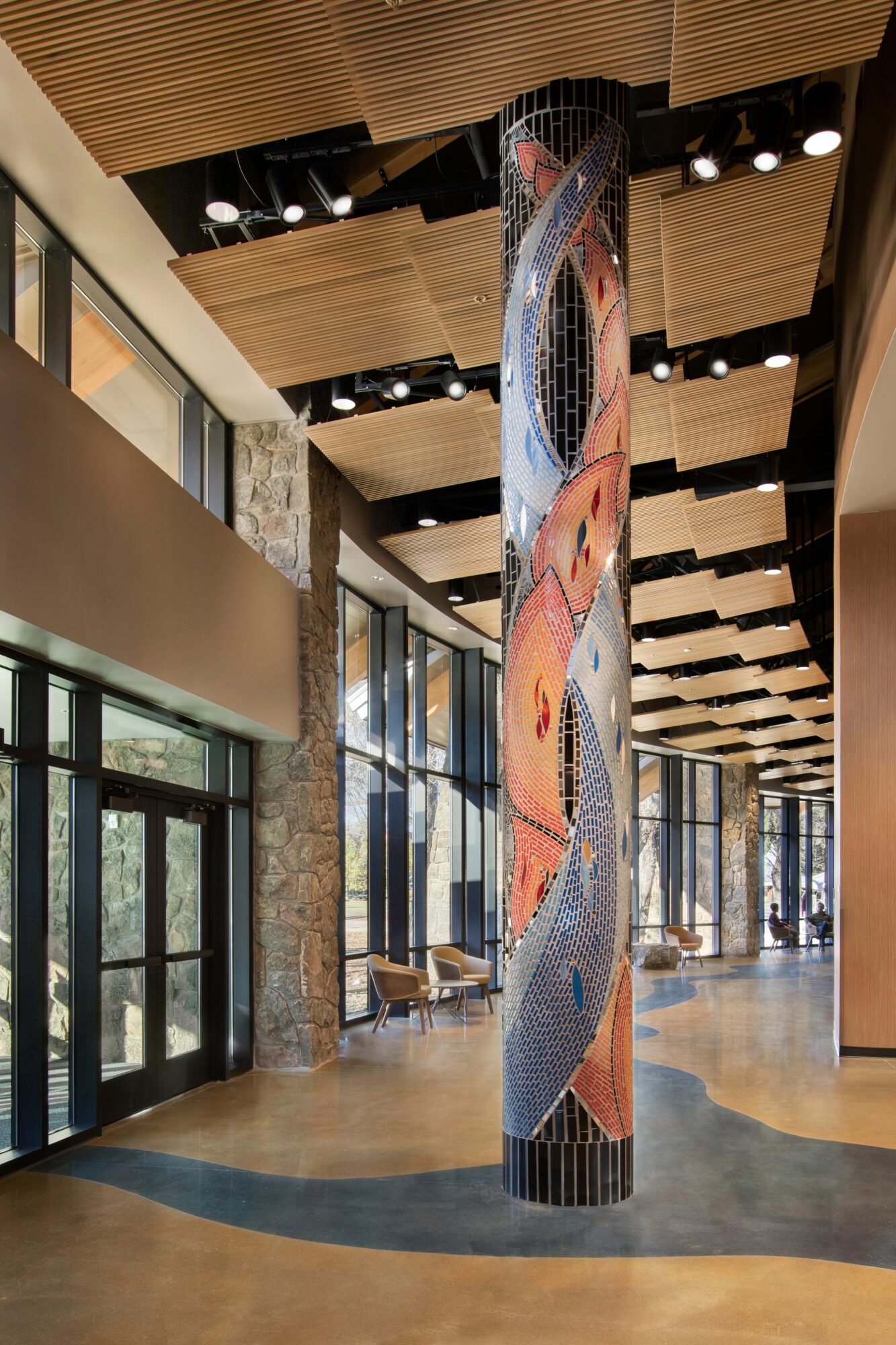 Tall cylindrical mosaic sculpture in a modern interior with wooden ceiling panels and large glass windows, reflecting natural light.