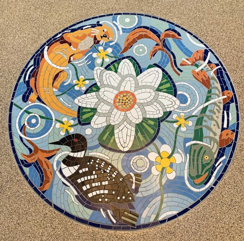 Colorful circular mosaic with fish, flowers, and a large white flower in the center, on a sandy background.