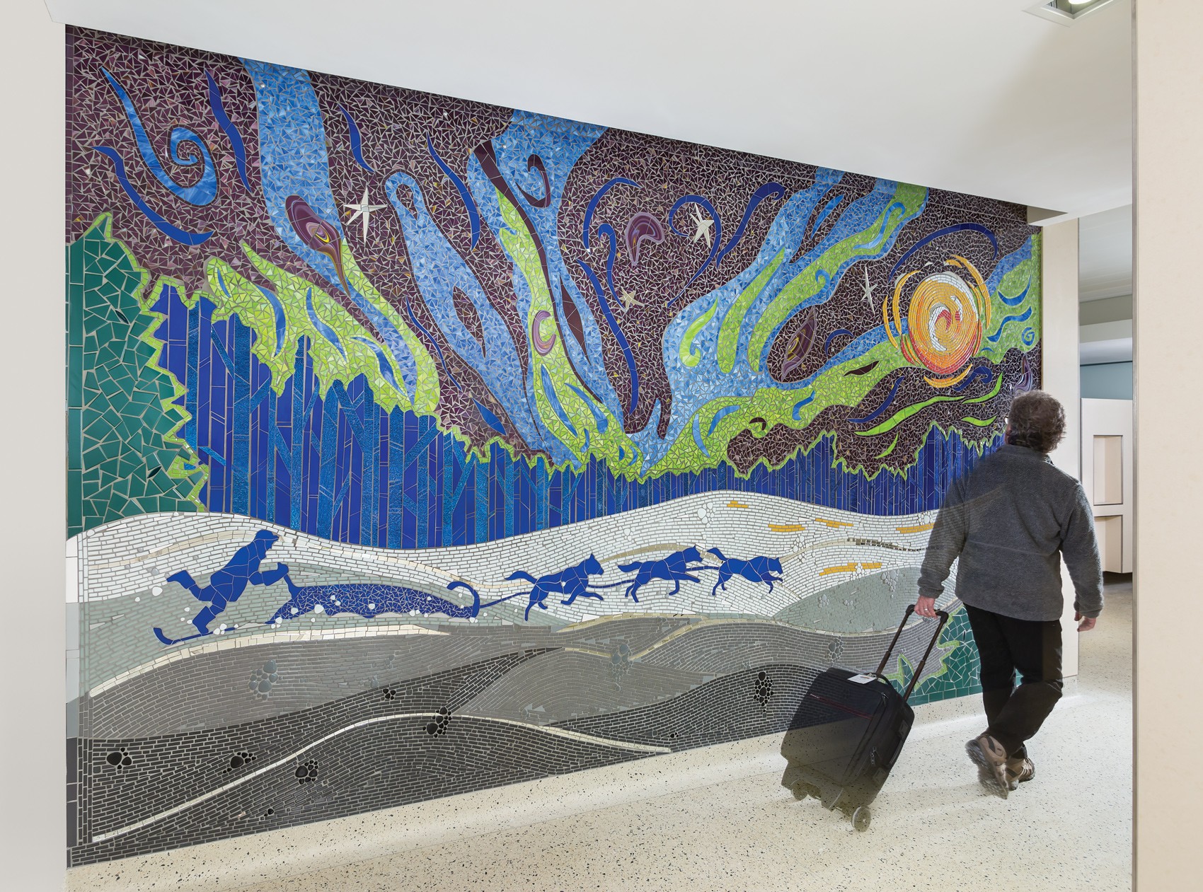 Colorful mosaic mural depicting a night sky, mountains, and animals, with a person walking and pulling a suitcase in an indoor space.