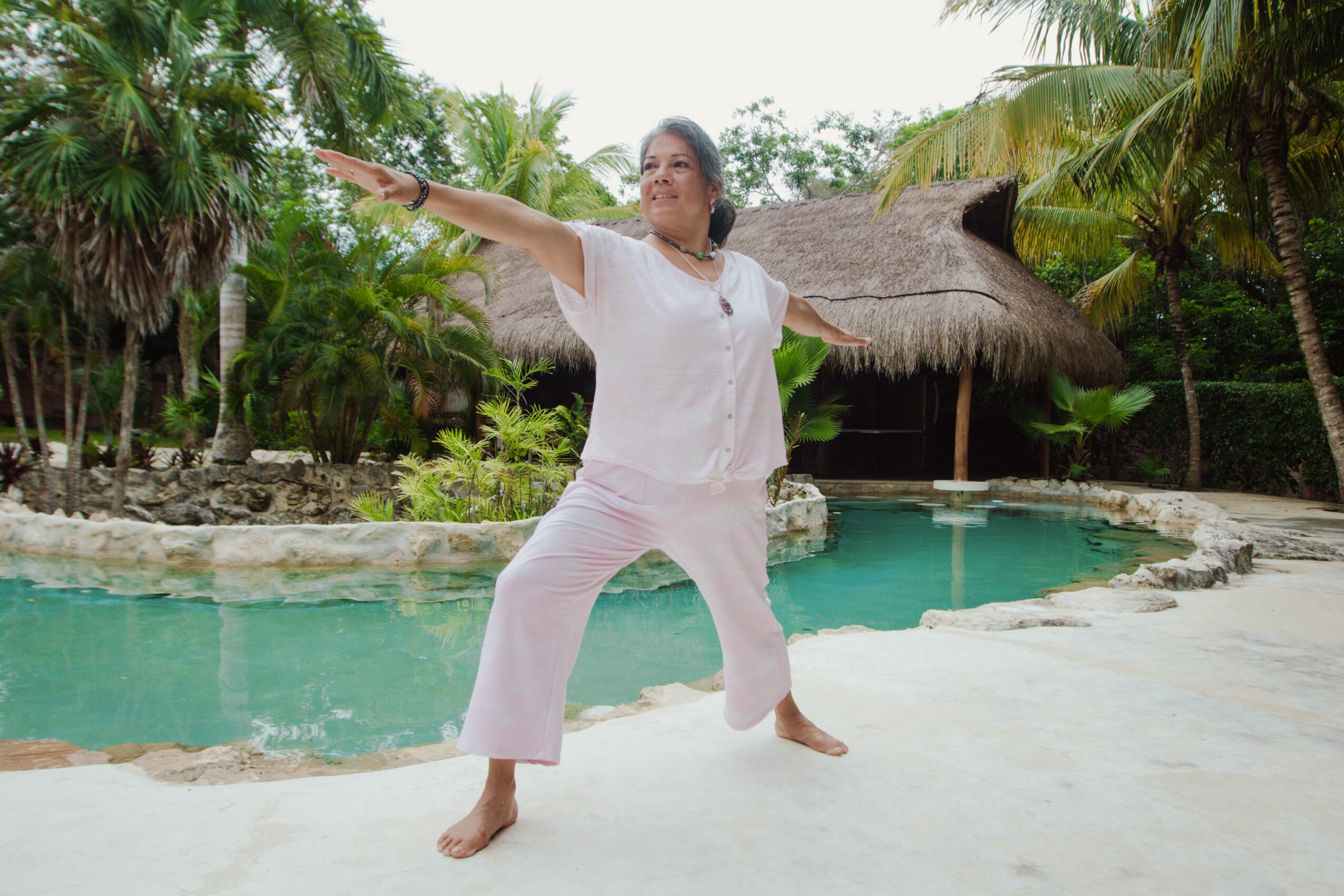 Woman in white pants and top standing on a beach near a pool, with arms outstretched, surrounded by palm trees and thatched hut.