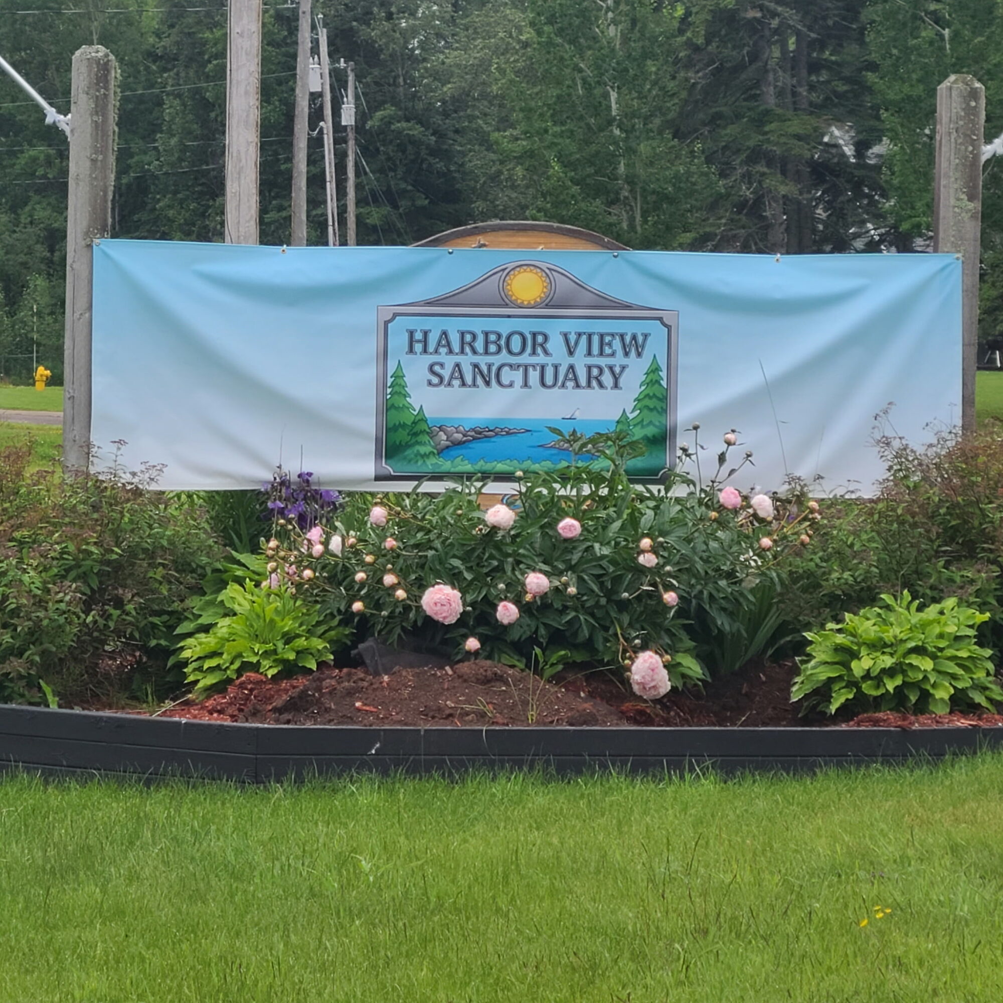 Sign for Harbor View Sanctuary with flowers and greenery in front, trees and utility poles in background.