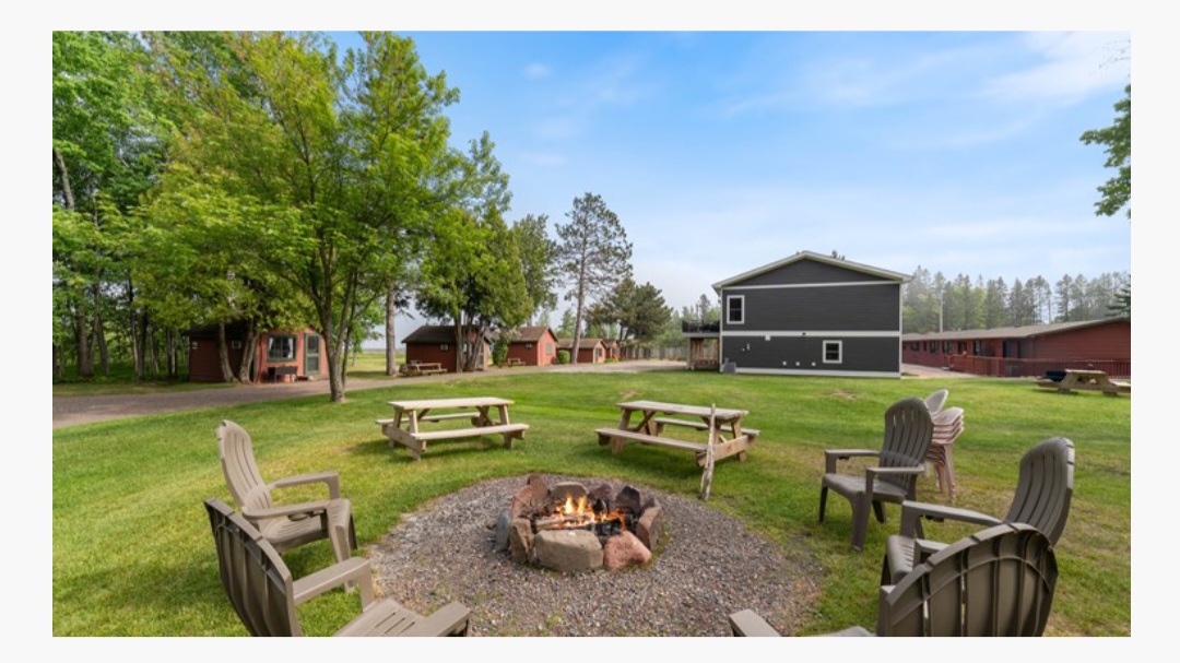 Outdoor scene with chairs around a fire pit, green grass, trees, and a black building in the background.