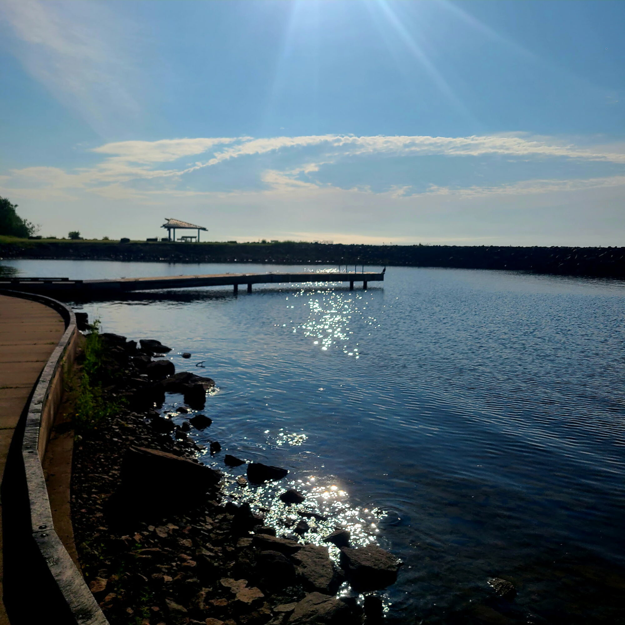 Calm water with a dock, a walkway on the left, and a sky with clouds and sunlight reflection.