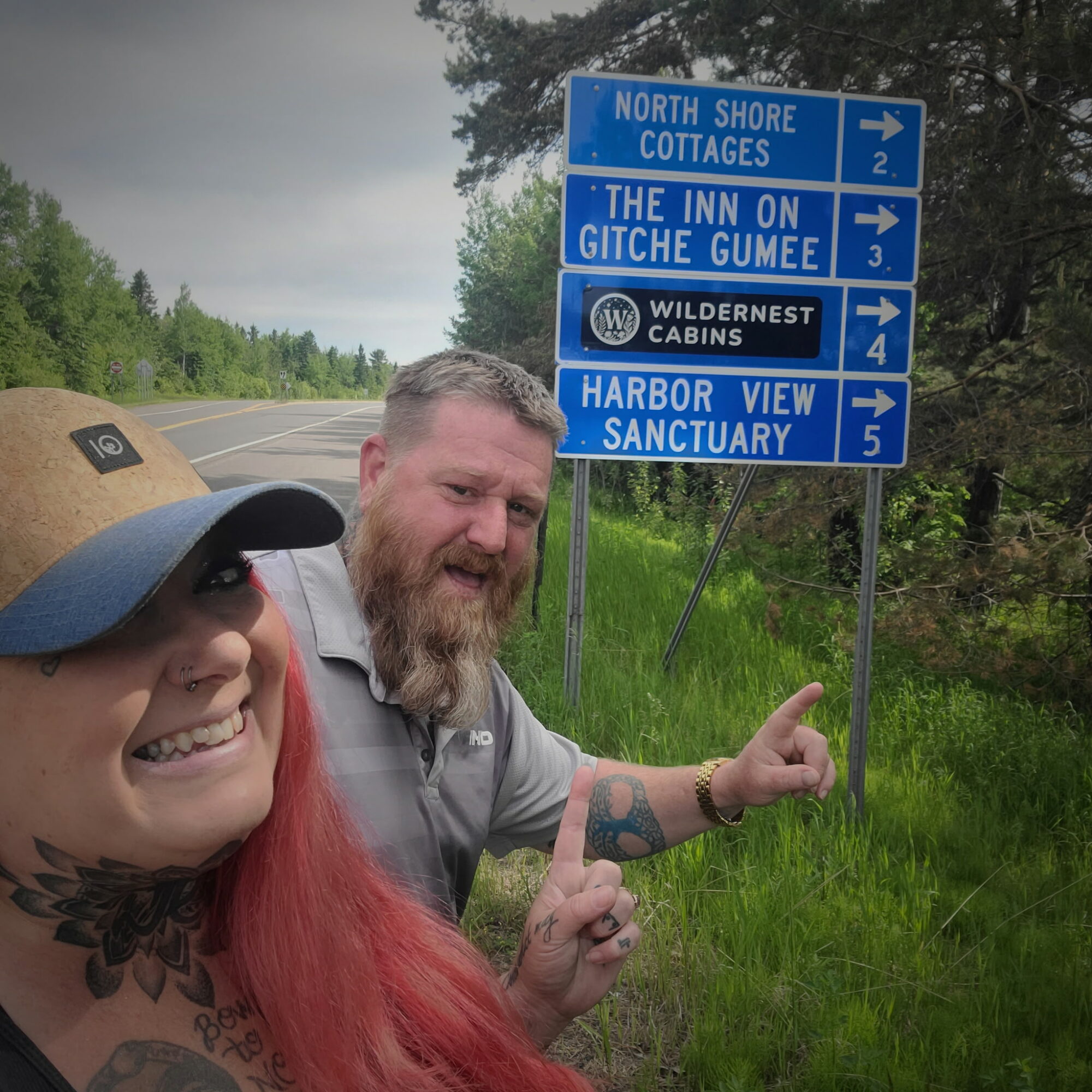 A smiling woman with red hair and a tattoo, and a bearded man, pointing at a blue road sign on a grassy roadside.