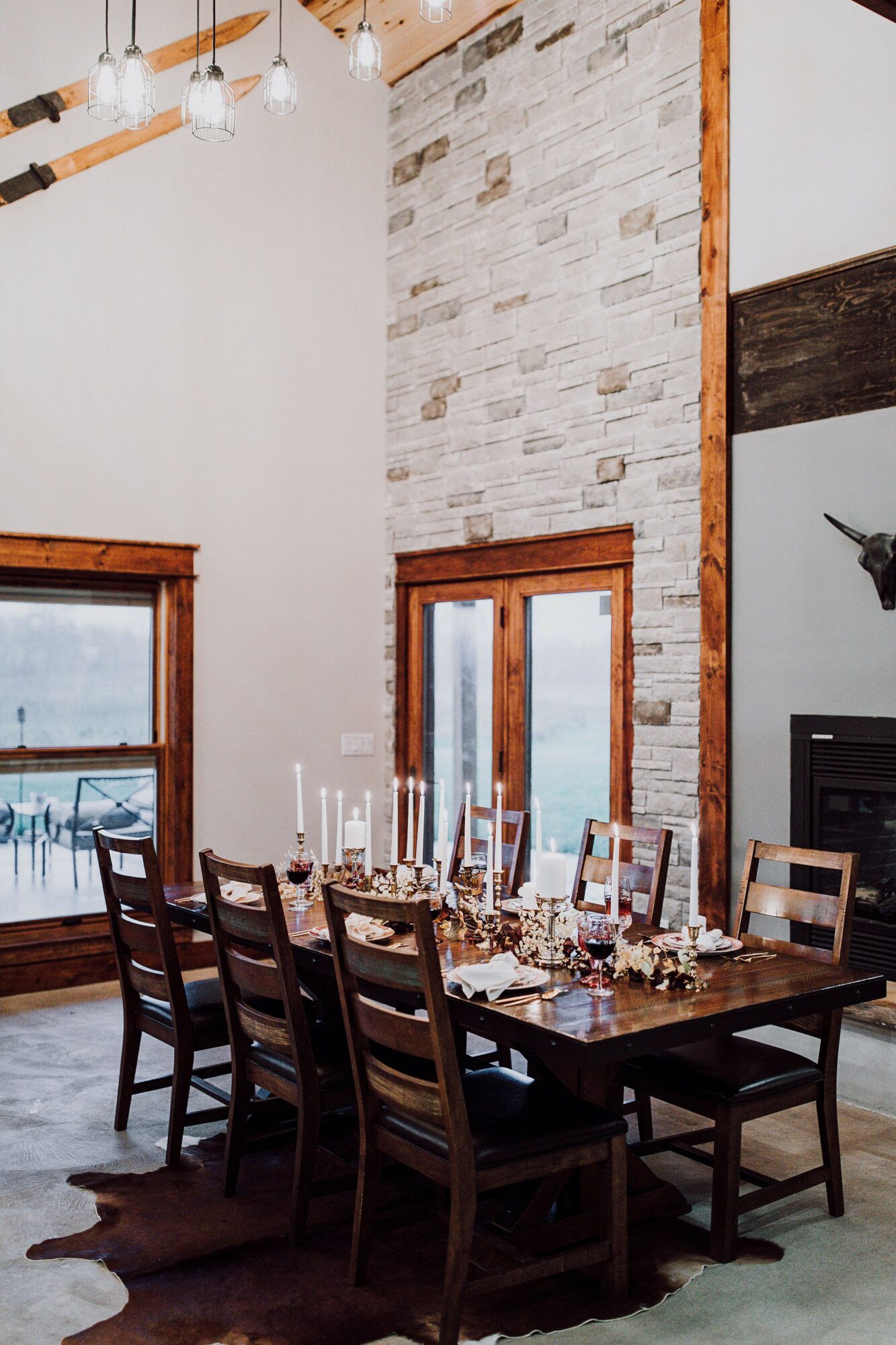 Dining table with chairs, candles, and tableware in a room with large windows, brick wall, and wooden accents.