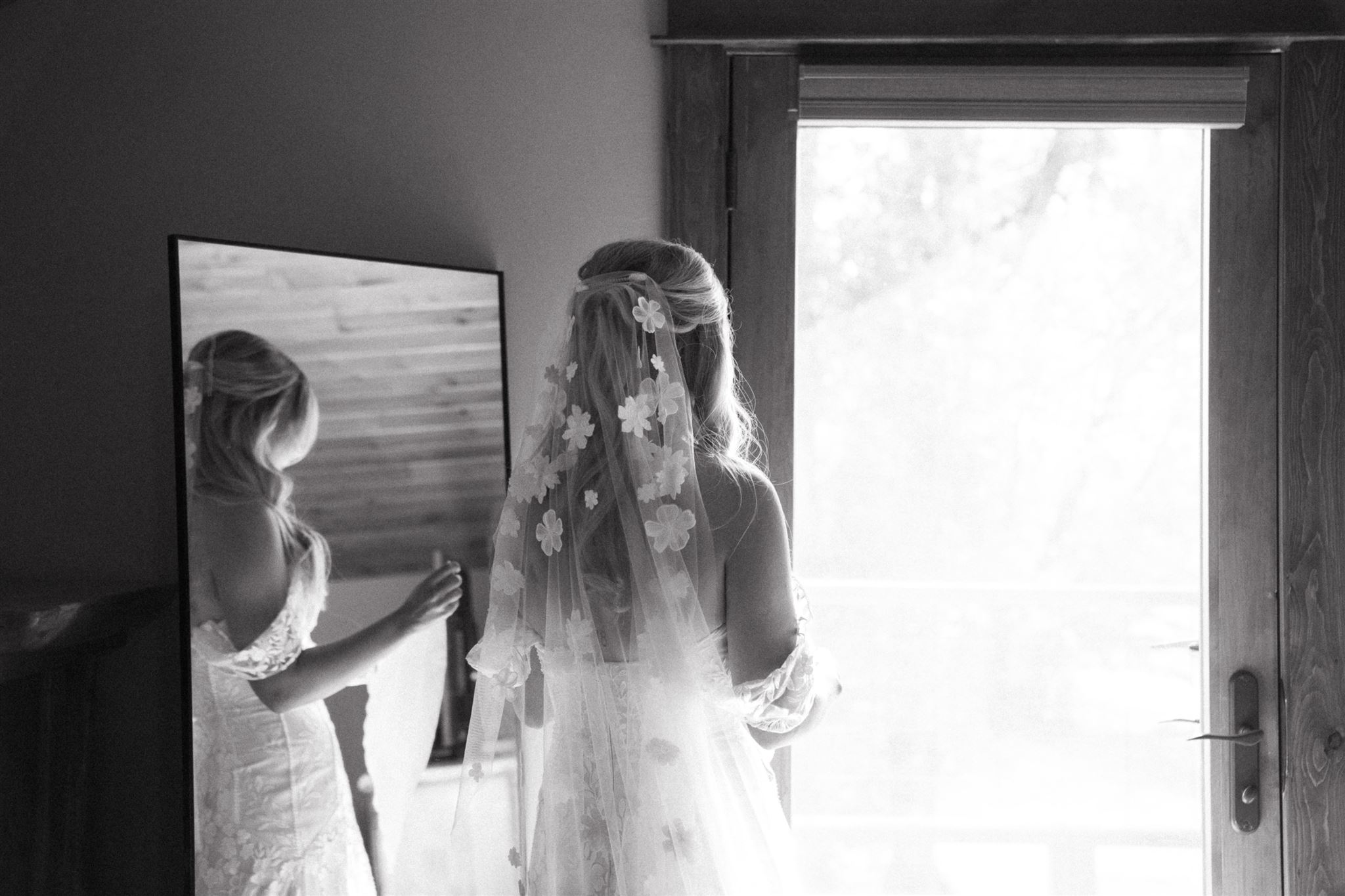 Bride with veil looking at her reflection in a mirror near a window, bright light outside, wooden door frame visible.
