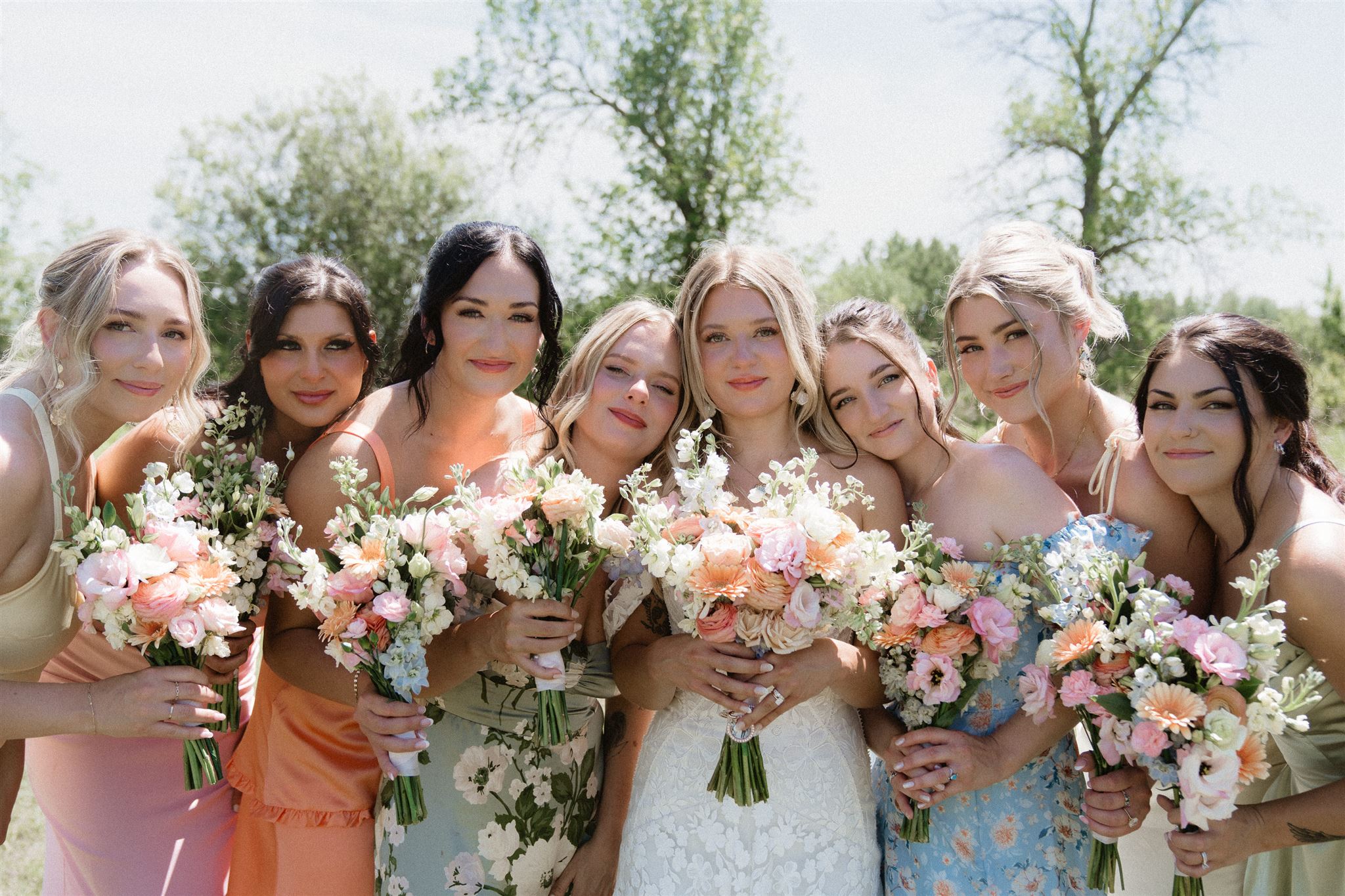 Group of eight women holding bouquets of flowers outdoors, smiling, with trees and sky in background.
