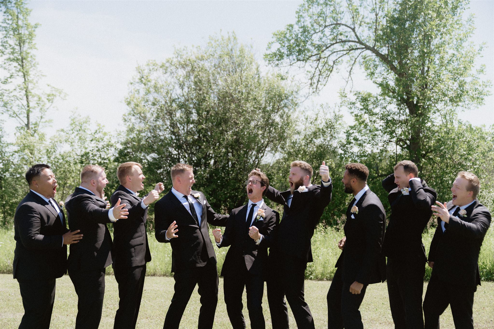 Group of men in suits standing outdoors, some holding drinks, trees and sky in background.