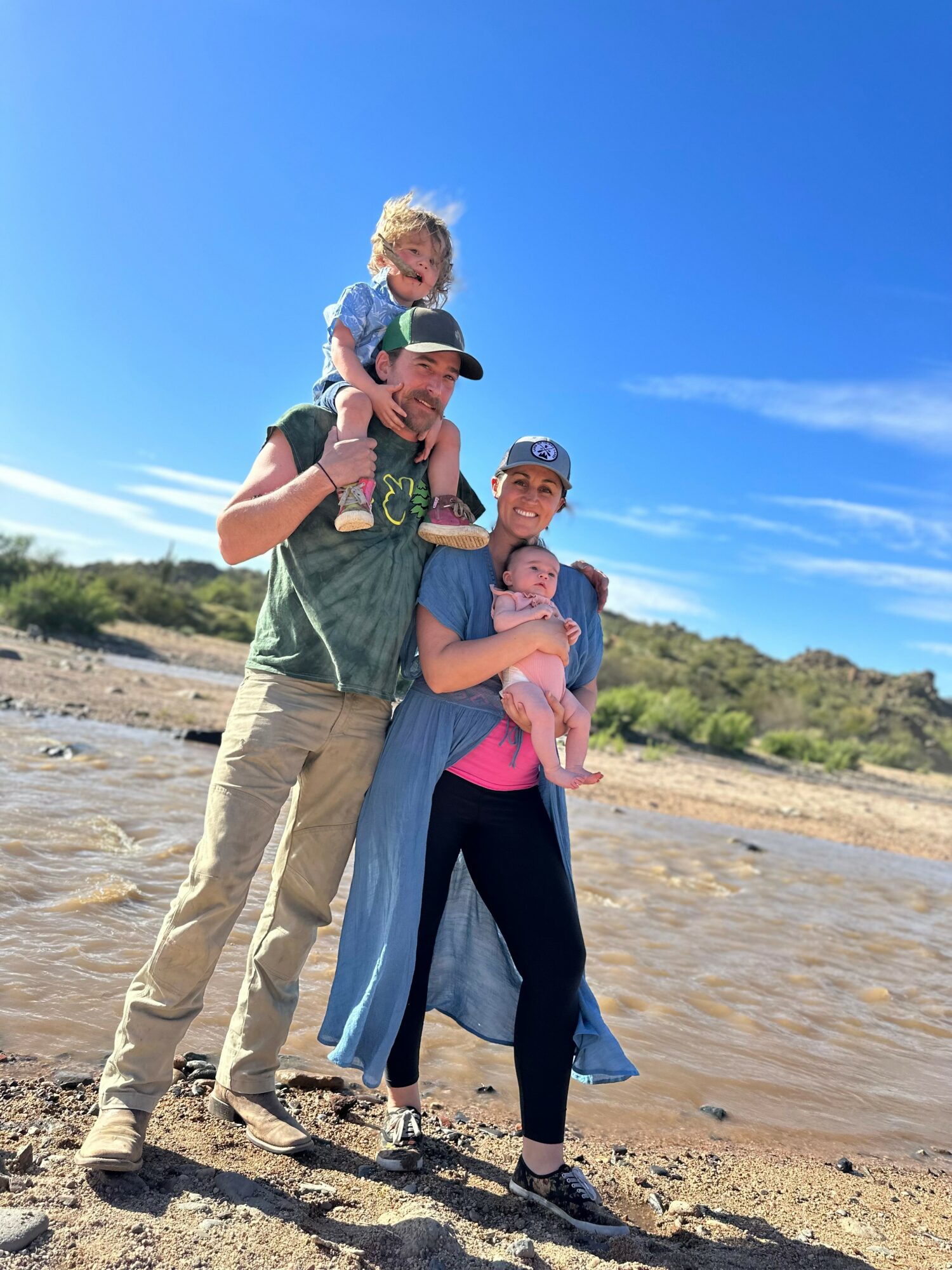 Family of three outdoors on a sunny day, with a child on the father's shoulders and a woman holding a baby, standing on a sandy area near water.