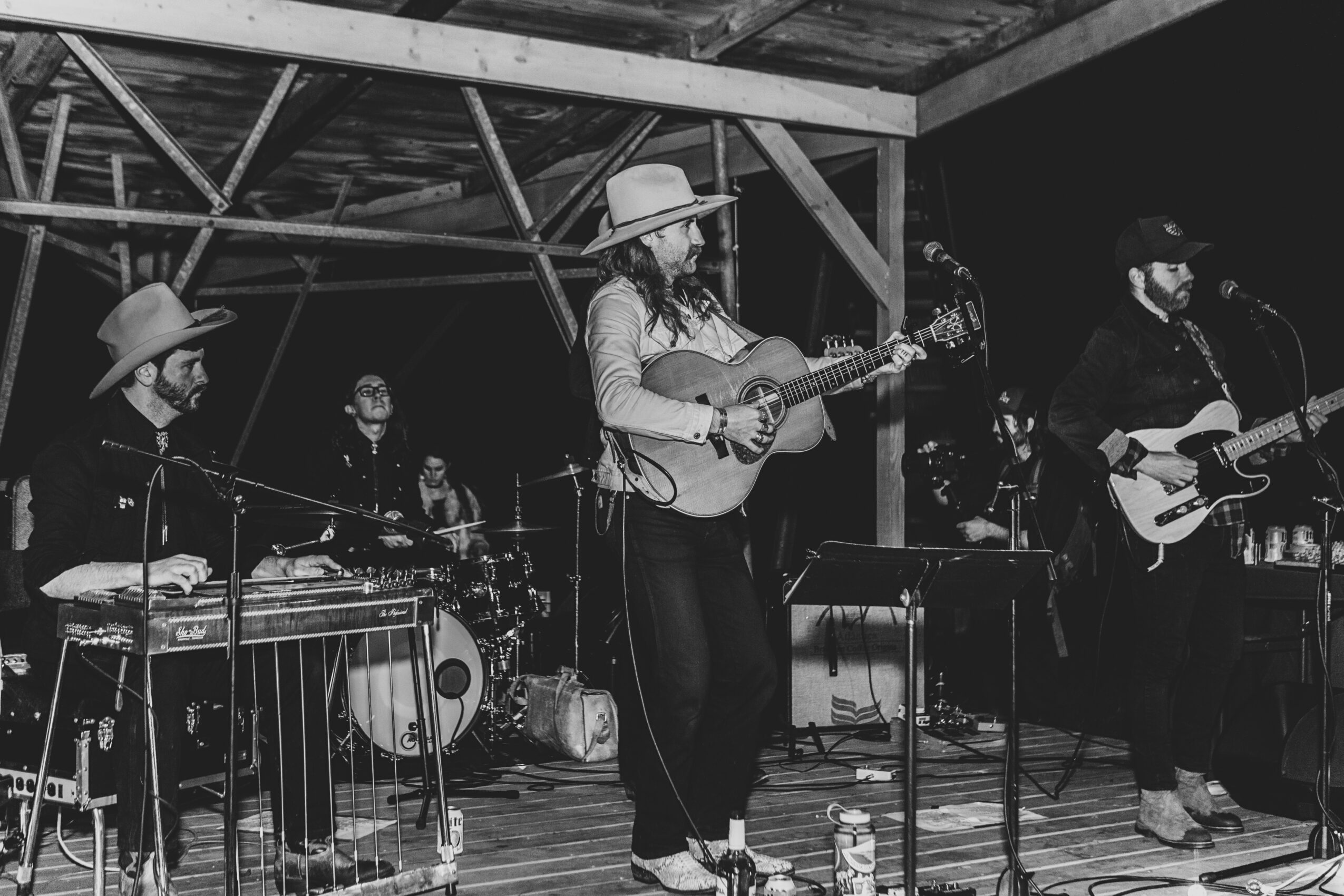 Group of five musicians performing on stage at night, playing guitars, drums, and percussion, under a wooden structure, black and white photo.
