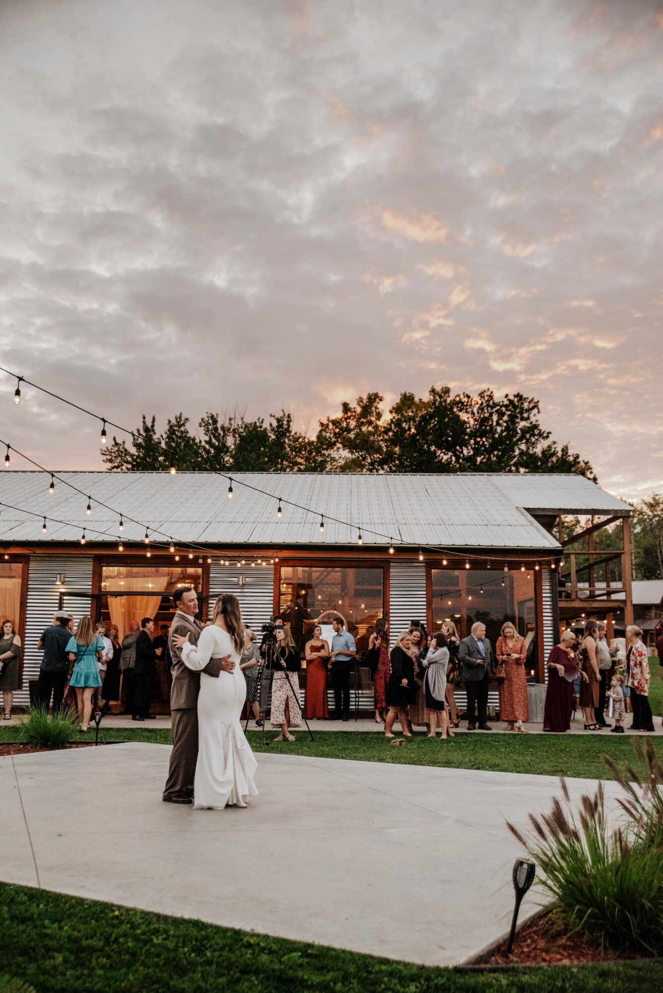 Couple dancing outdoors at sunset with string lights and a gathering of people in the background.