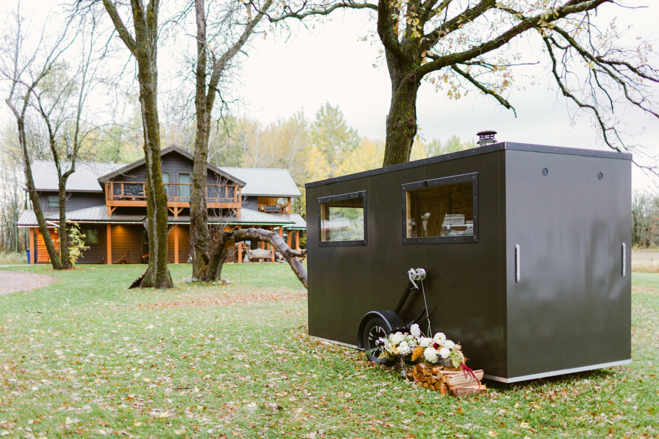 Black food truck with flowers and a basket in front, trees and a house in the background, outdoors in a park-like setting.