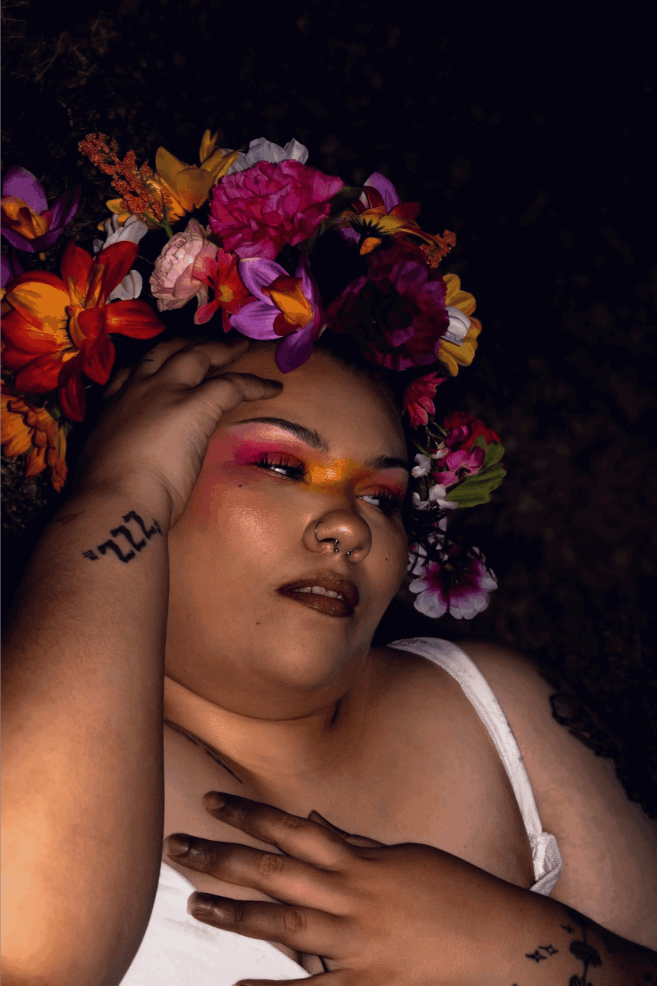 A woman with a floral crown, makeup, and tattoos, lying with her eyes closed and hand on her face.