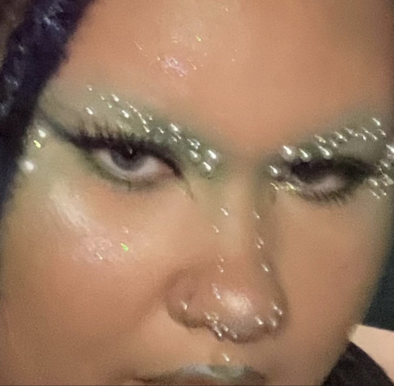 Close-up of a person's face with glittery makeup and a nose ring, eyes partially closed, and a serious expression.