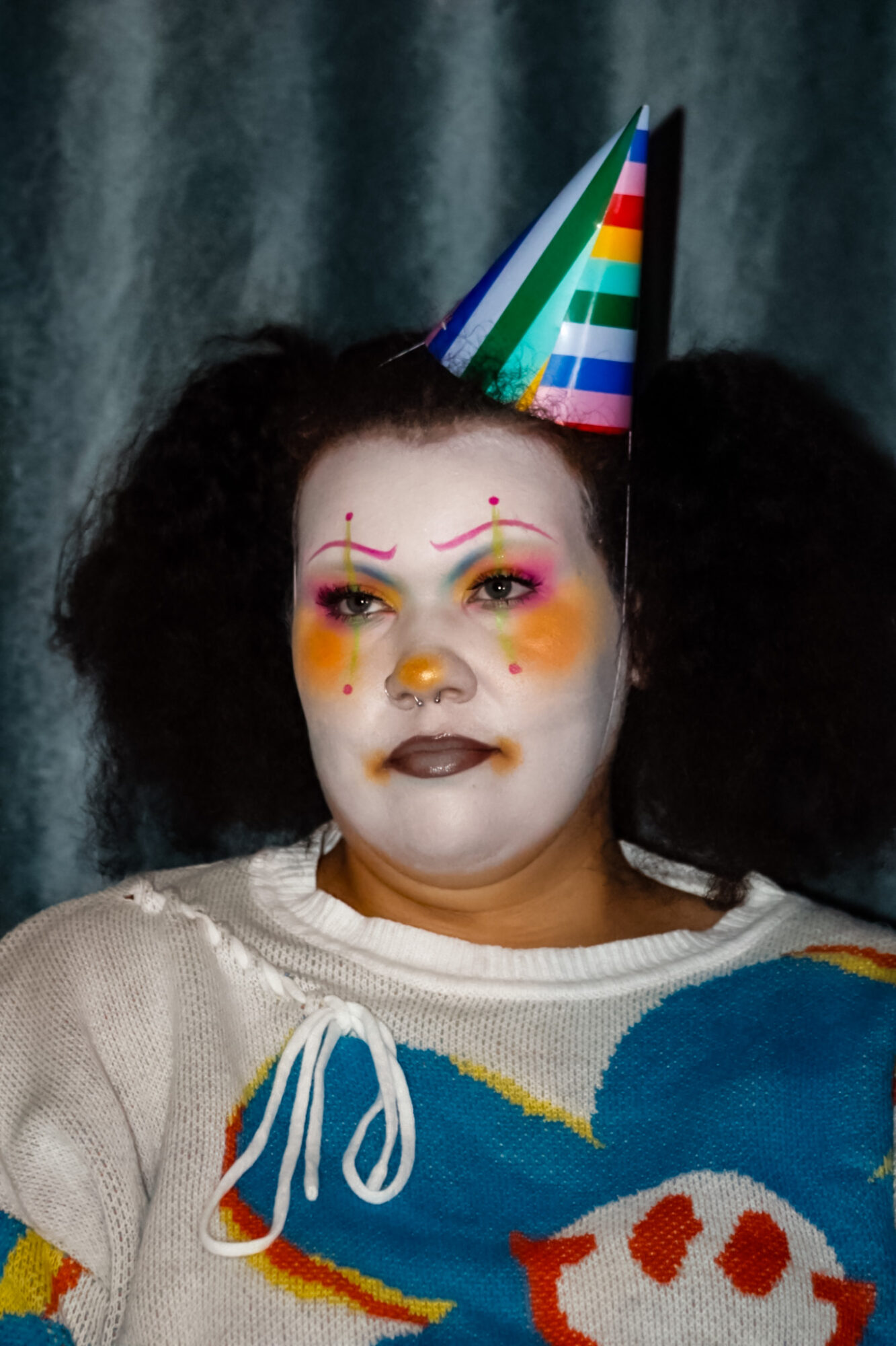 Child with clown makeup, rainbow hat, and colorful clothing, celebrating a party, against a dark background.