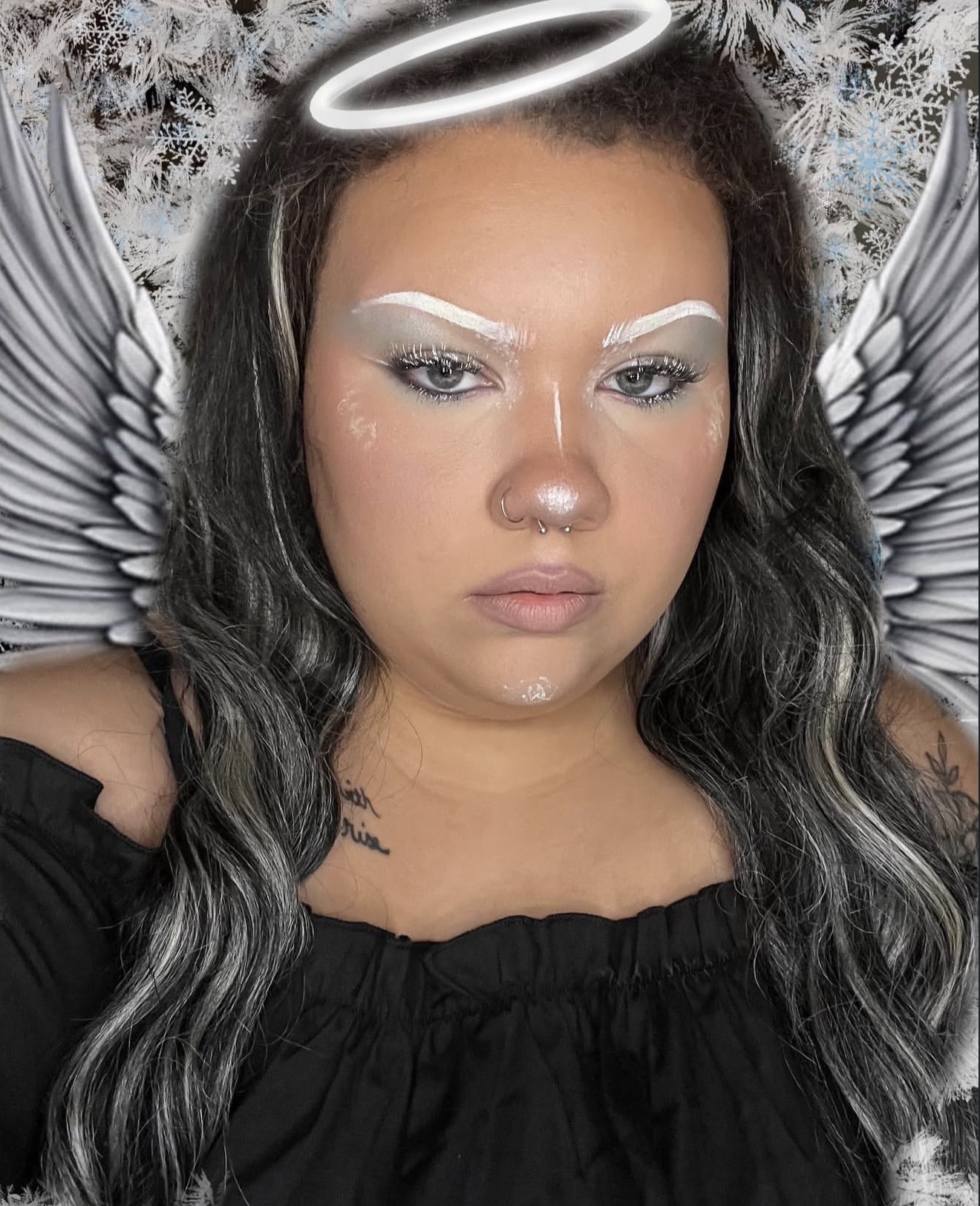 Person with angel wings, halo, and makeup, wearing a black top, with a snowy background, looking serious.