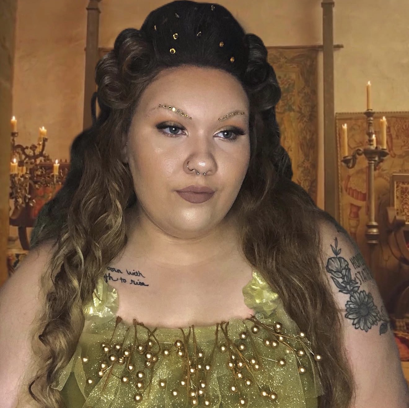 Woman with long wavy hair in a green dress with gold embellishments, tattoos on her arm, in a decorated room with candles