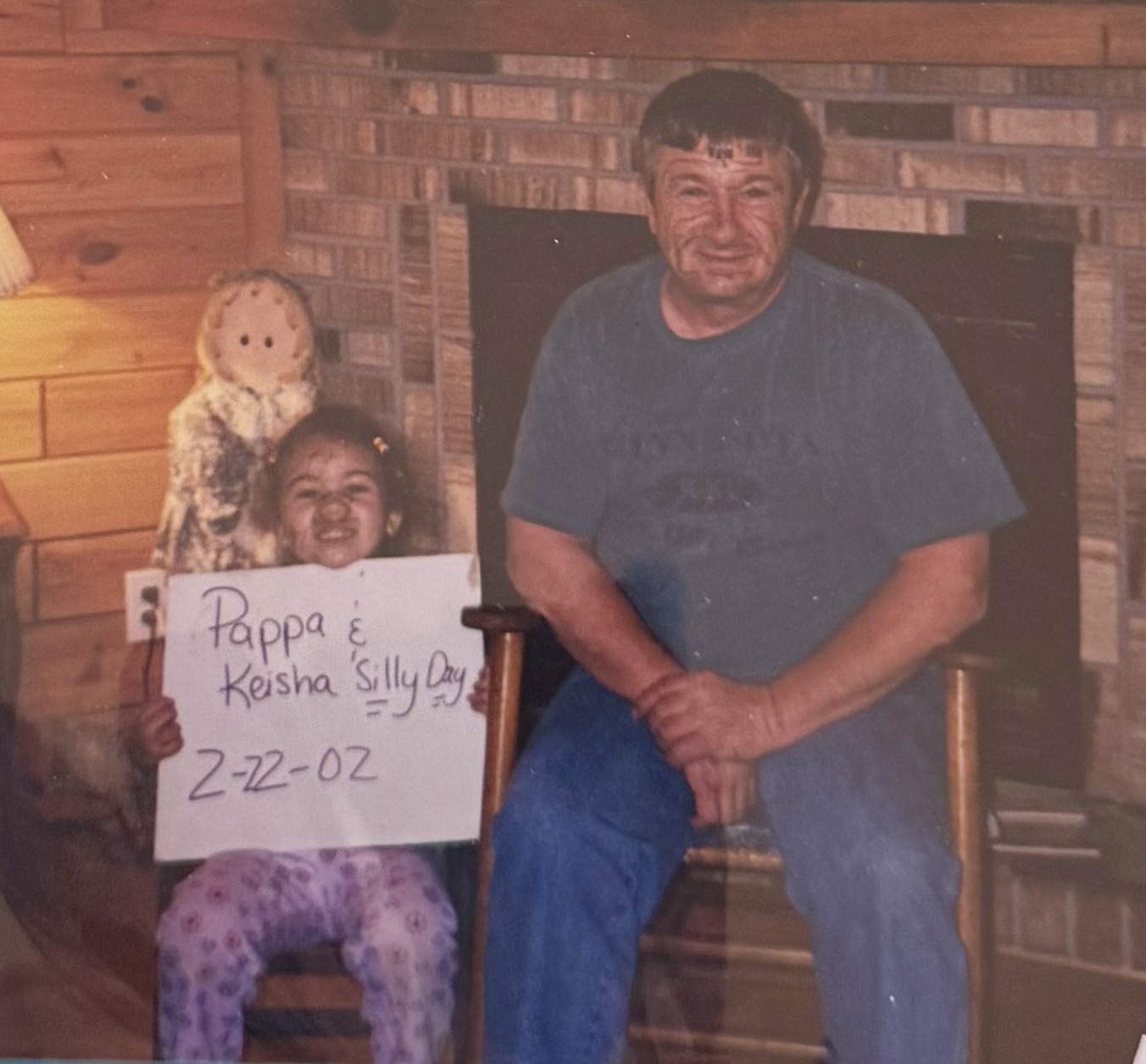 A man and a young girl sitting in front of a brick fireplace, holding a sign with handwritten text, smiling.