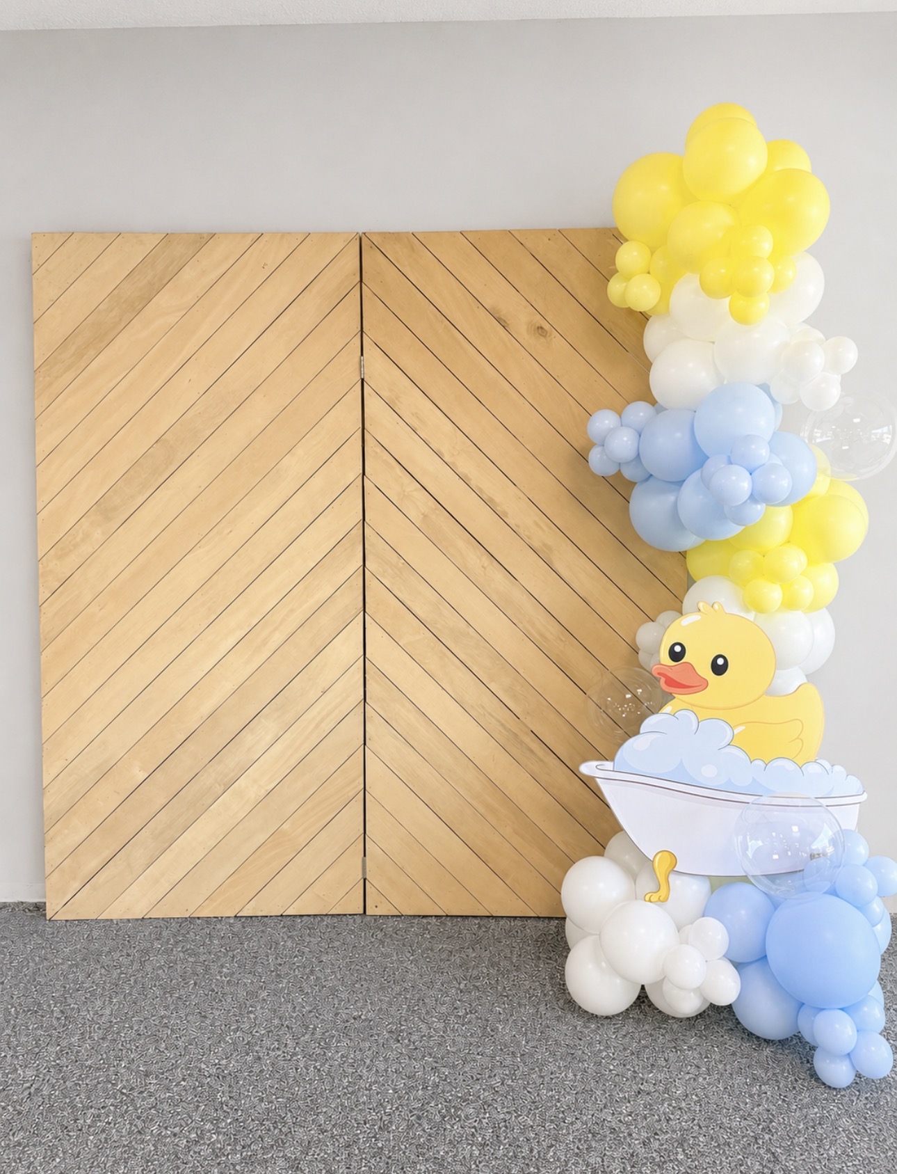 Decorative arrangement of yellow, blue, and white balloons with a rubber duck, positioned beside a wooden wall, on a gray floor.