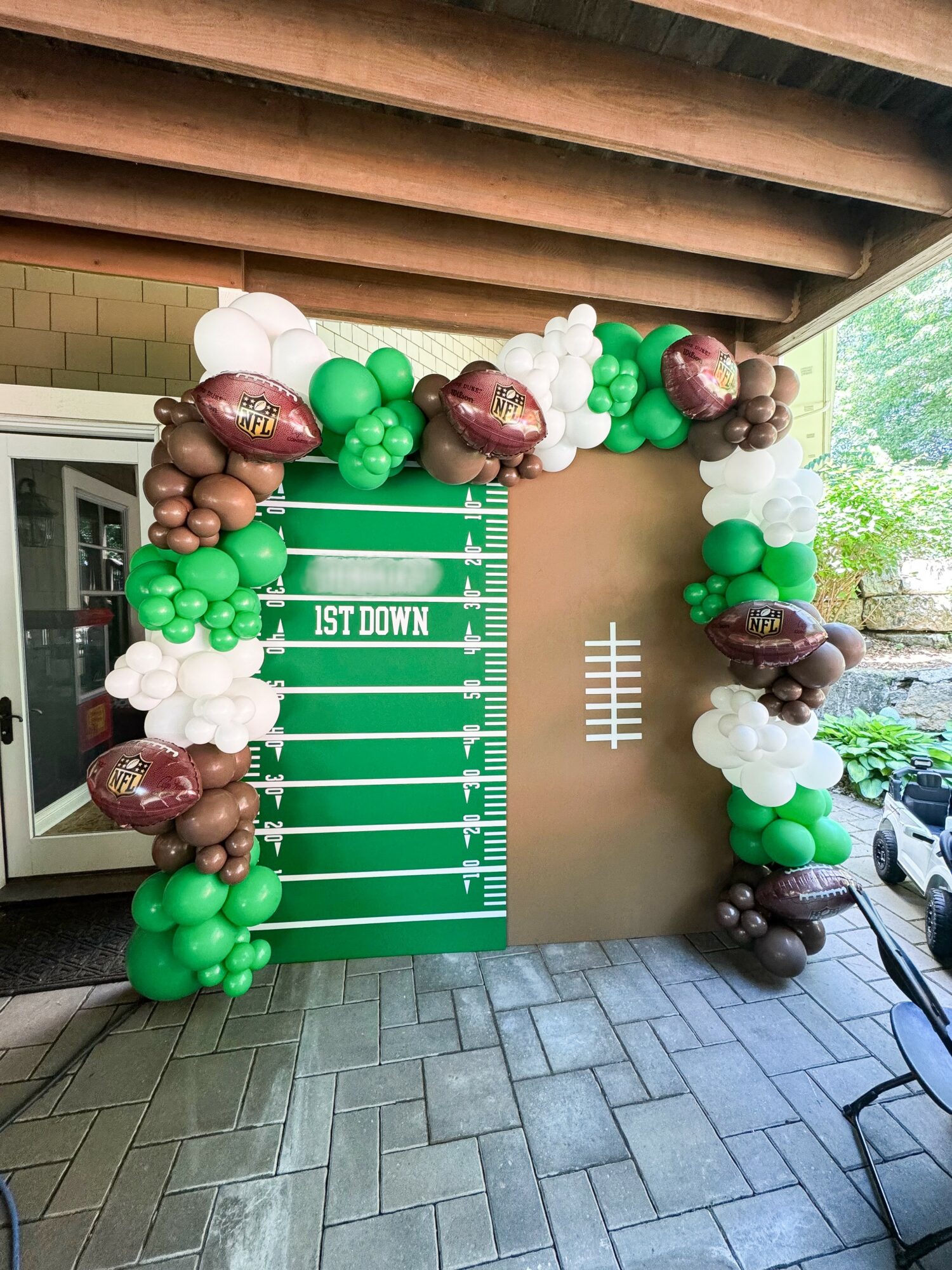 Balloon arch with green, white, brown, and purple balloons surrounds a green football field backdrop with yard lines, on a patio.