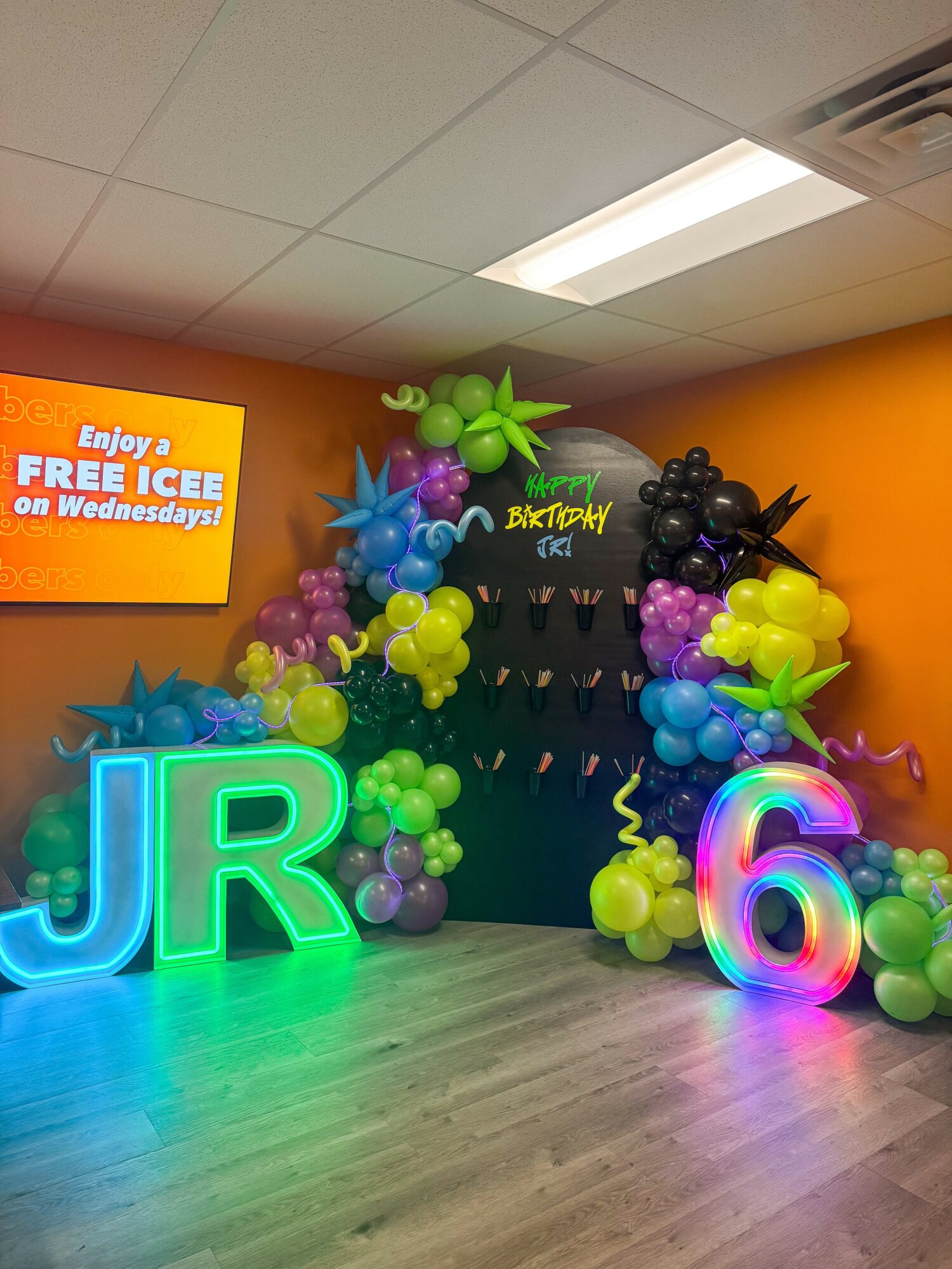 Colorful balloon arch with large illuminated letters 'JR' and the number '6' in front, inside a room with orange walls and a ceiling light.