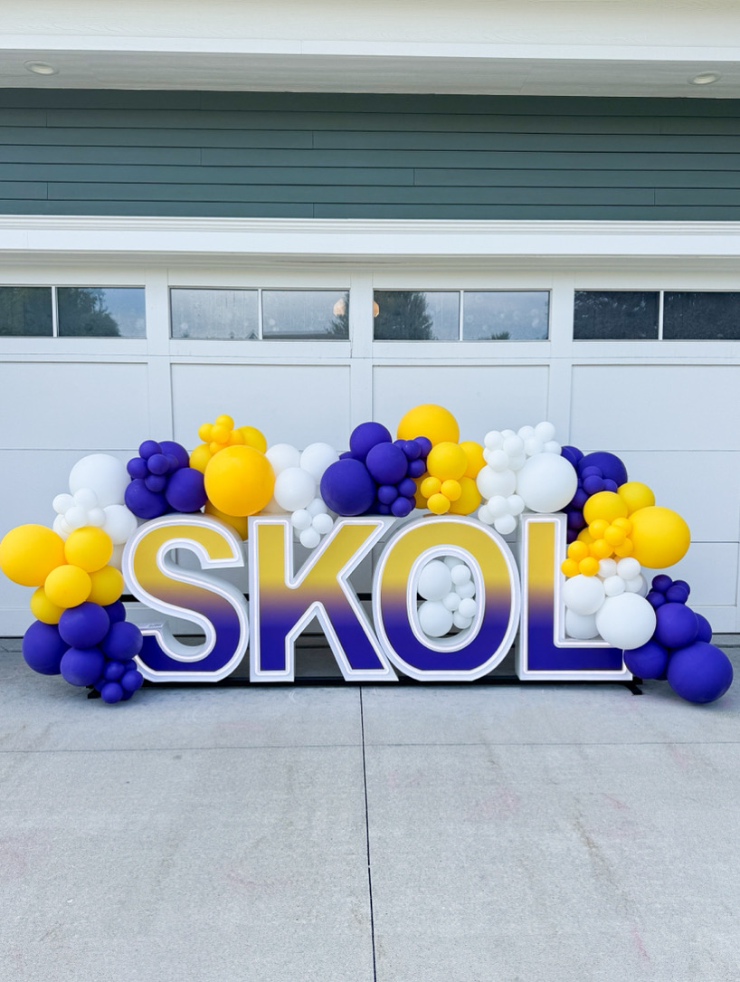 Large colorful balloons in yellow, purple, and white surround the word SKOL in front of a garage door.