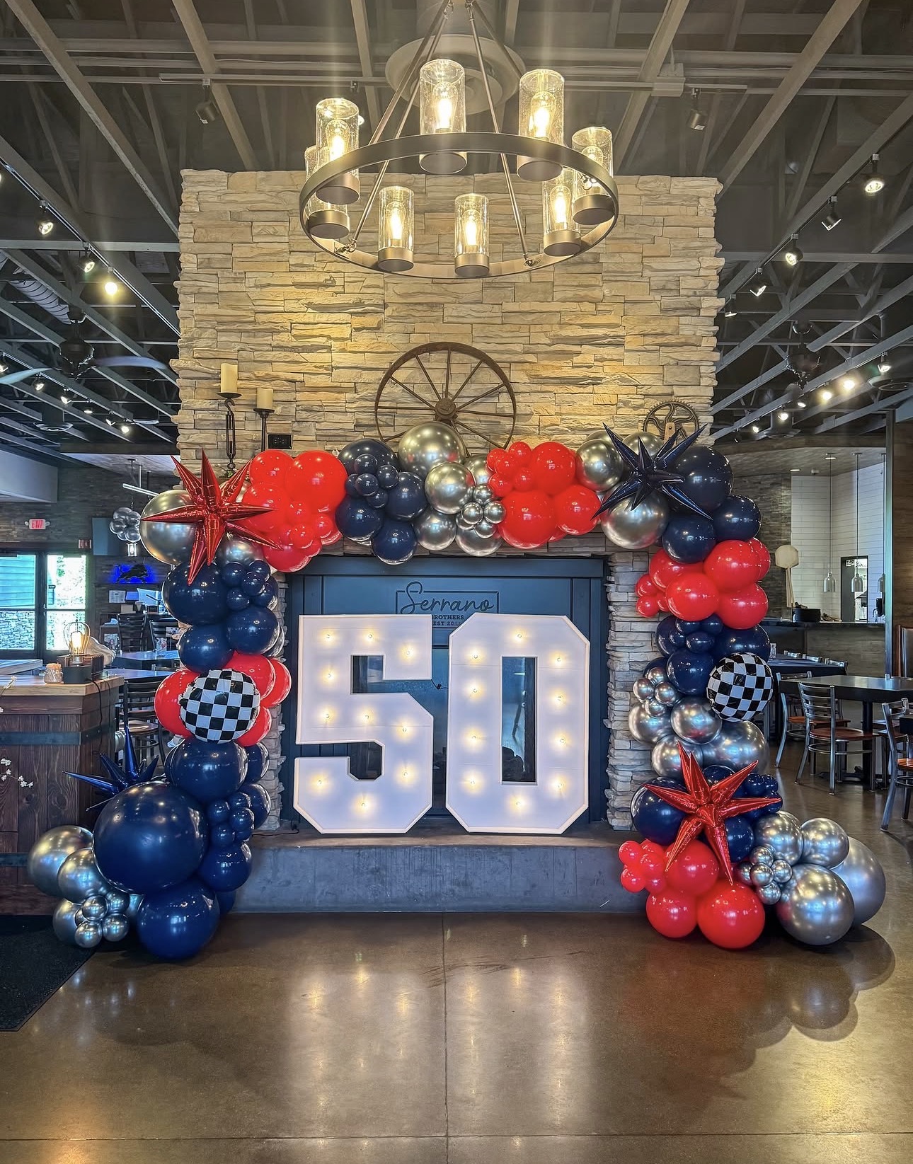 Decorative 50th birthday display with balloons and lights in a restaurant setting, featuring a large illuminated number 50, stone wall backdrop, and chandelier.