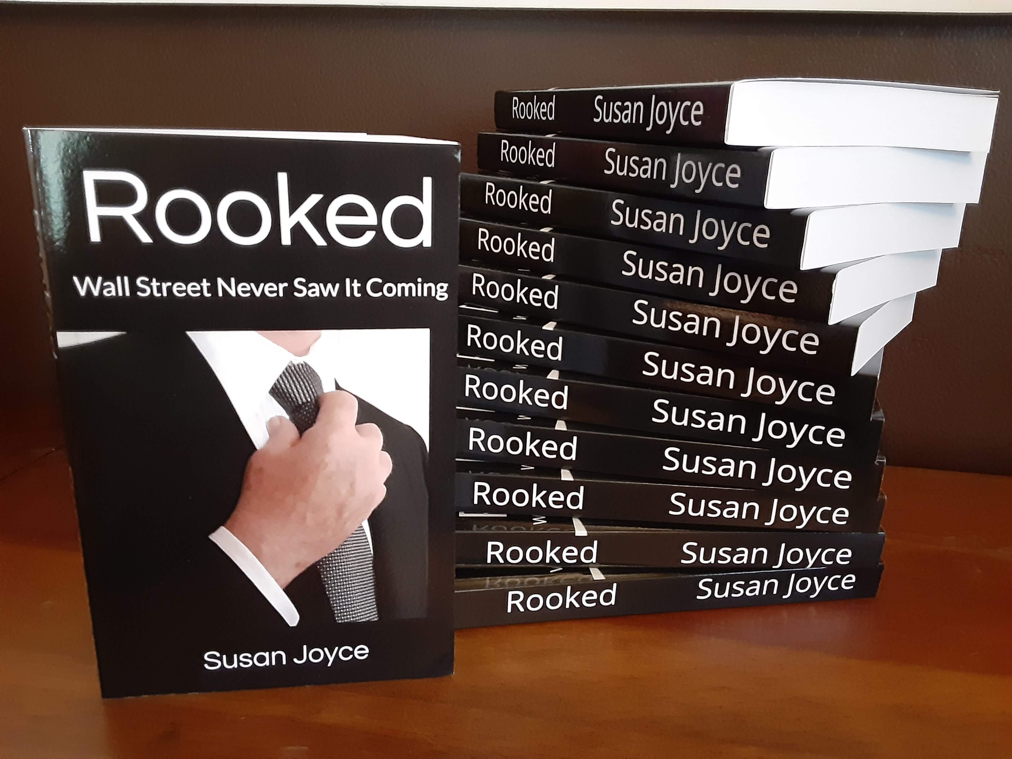 Book titled 'Rooked' by Susan Joyce with a hand holding a chess piece on the cover, stacked books behind, on a wooden surface.
