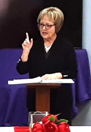 Woman with short blonde hair and glasses, gesturing with her right hand, sitting at a table with papers, in front of a purple backdrop.