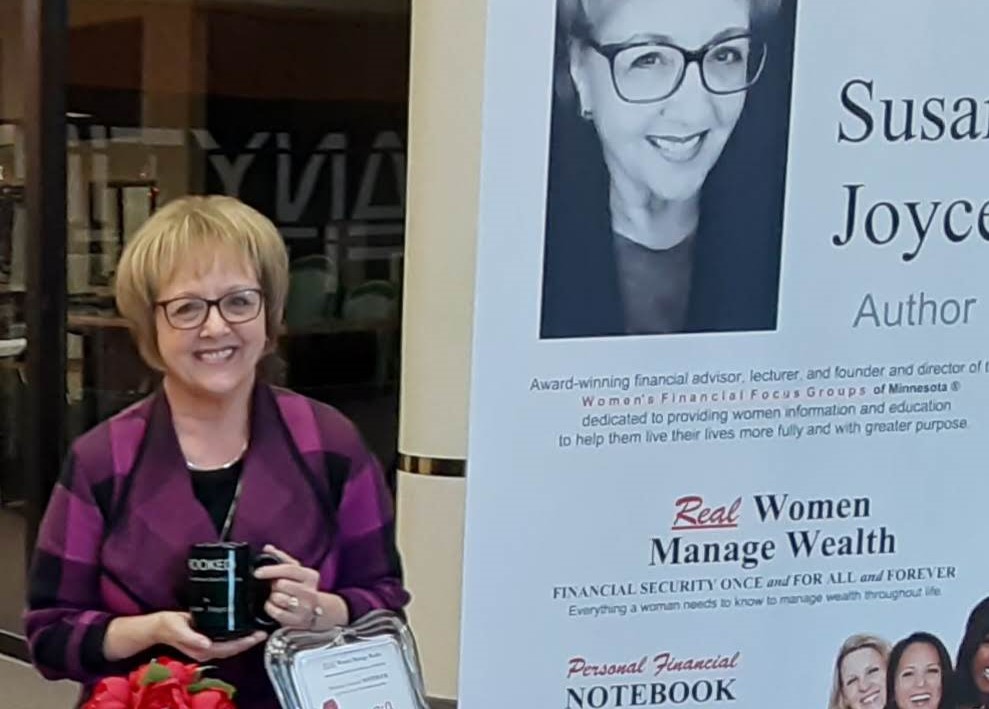 Woman with glasses holding a coffee mug and a framed certificate, standing next to a large poster of herself.