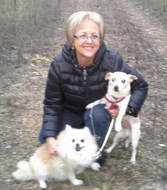 Woman with glasses crouching outdoors with two dogs on a dirt path, smiling at the camera.