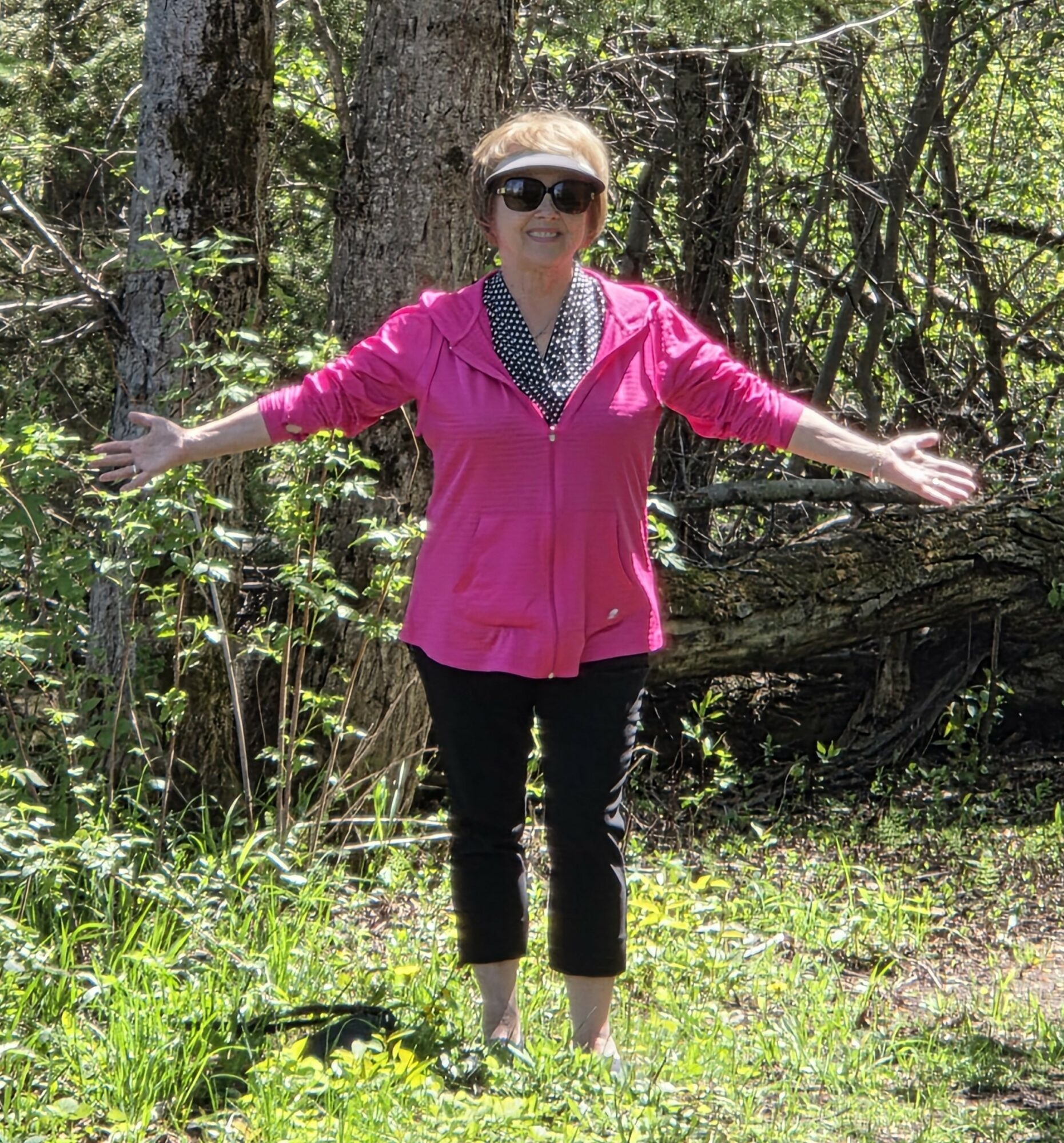 Woman standing outdoors with arms outstretched, wearing sunglasses, pink jacket, and black pants, surrounded by trees and greenery.