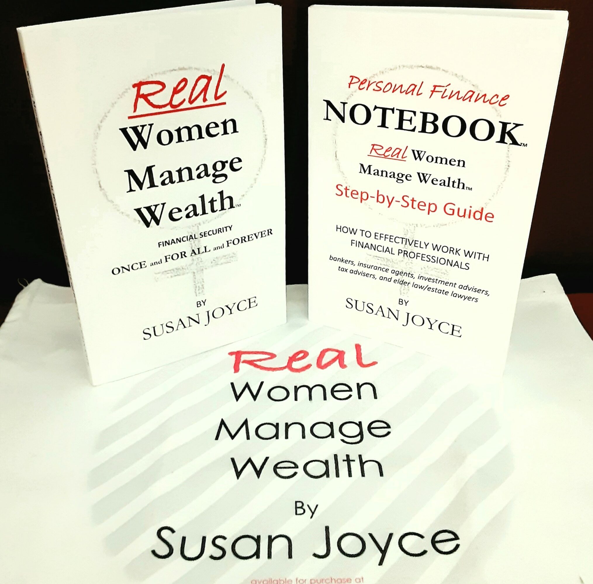 Three books titled 'Real Women Manage Wealth' and 'Personal Finance Notebook' by Susan Joyce, with one book open and two closed, on a white surface.