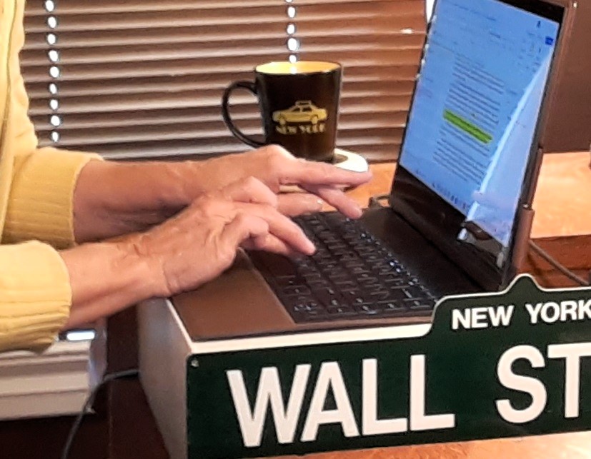 Person typing on a laptop with a coffee mug nearby, wall with blinds in the background.