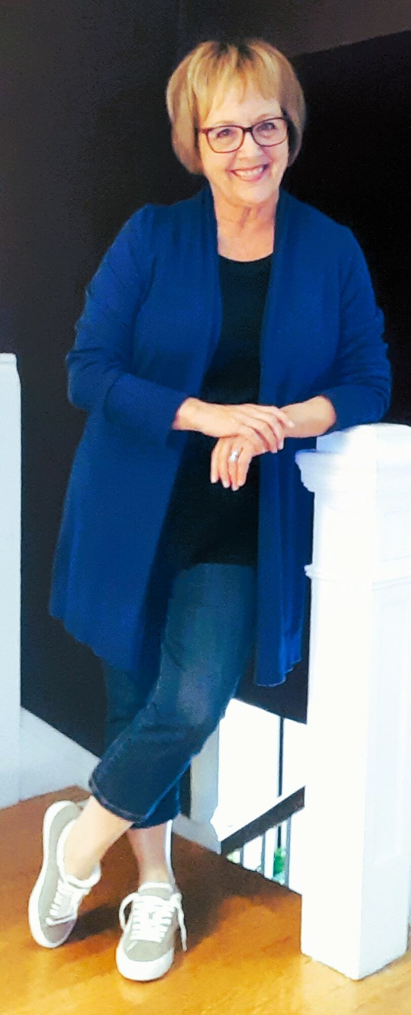 Woman with short blonde hair and glasses, wearing a blue cardigan and jeans, standing indoors near a white pillar.
