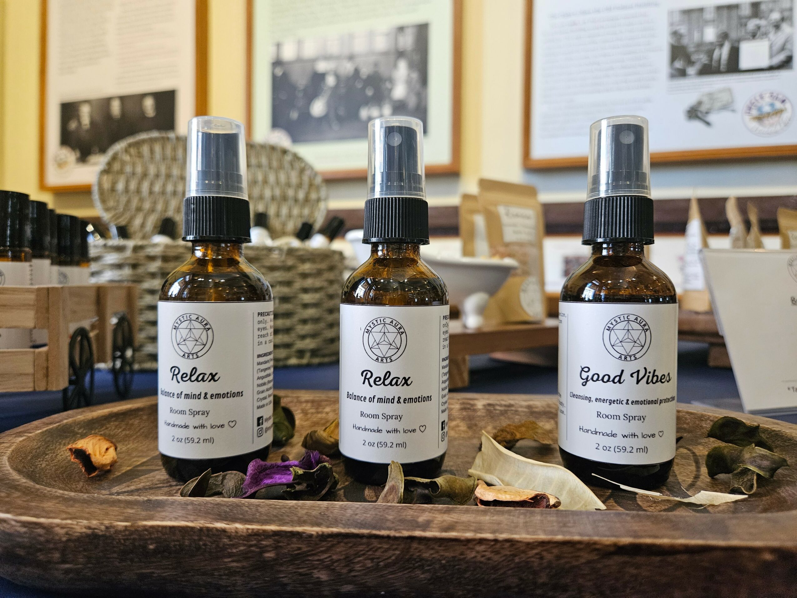 Three small bottles with spray tops on a wooden tray, surrounded by dried leaves, in a room with framed pictures on the wall.