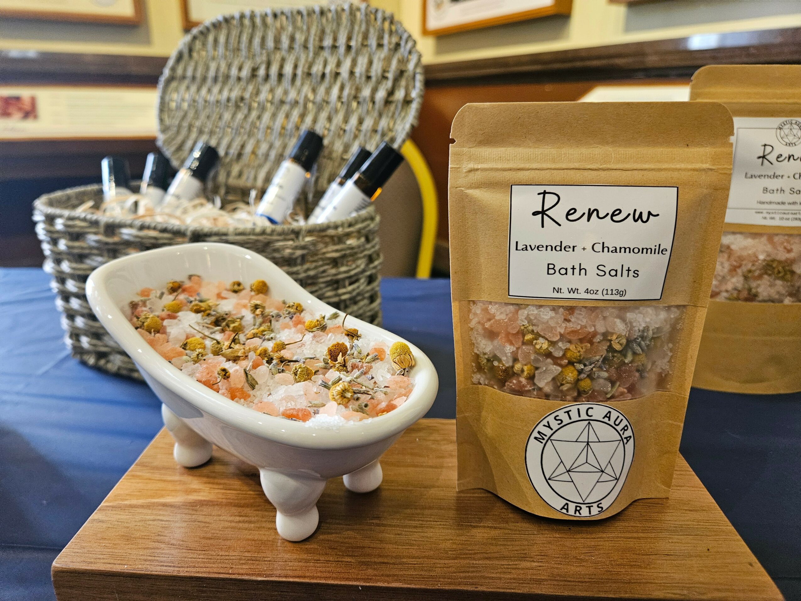 A small white dish with bath salts, a basket with pens, and two brown bags of bath salts on a wooden surface.