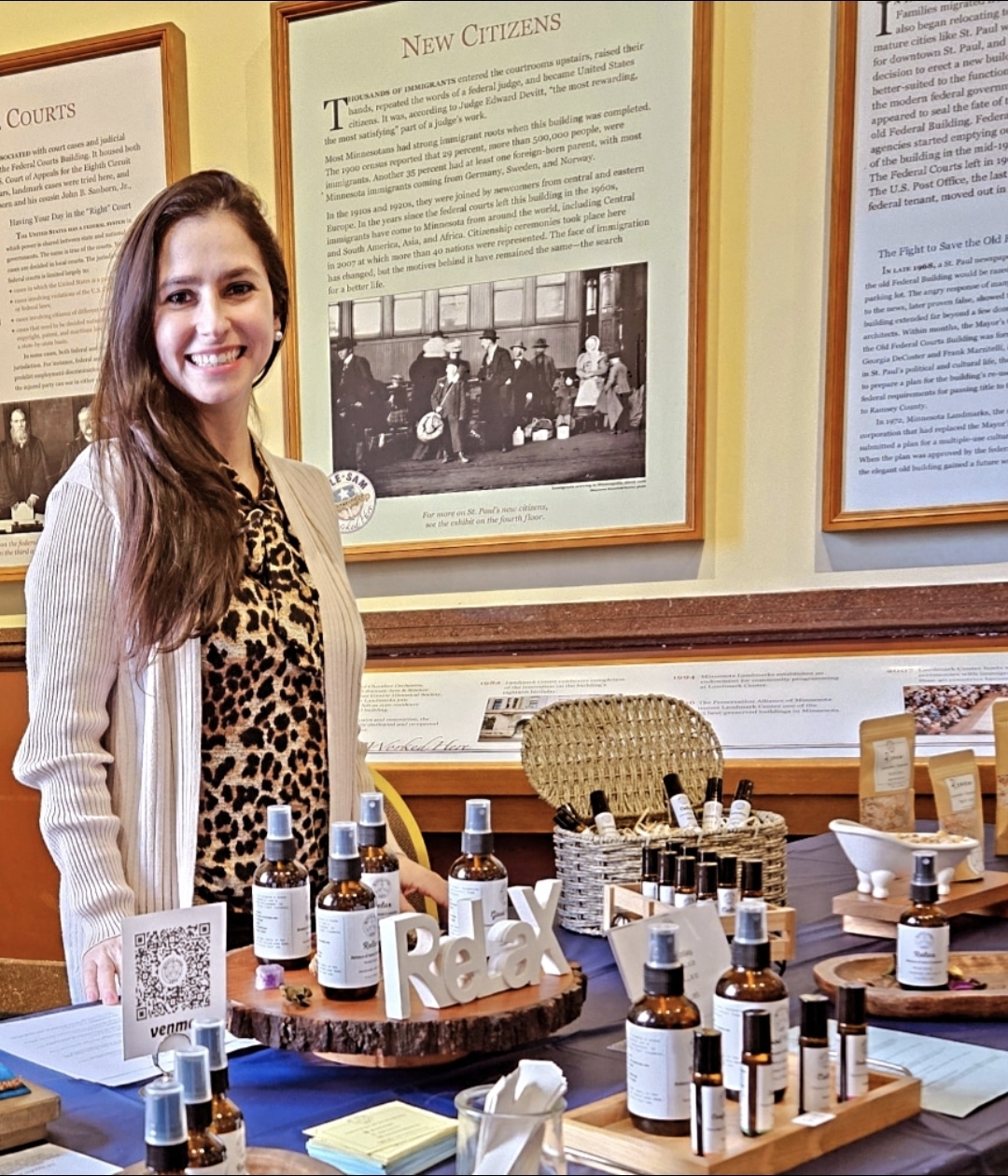 Smiling woman with long hair standing behind a table with bottles and skincare products in a museum or exhibit setting.