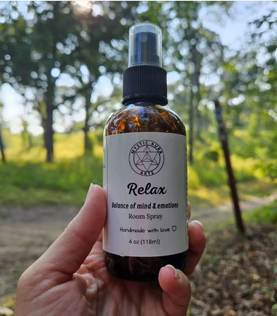 Hand holding a small amber spray bottle labeled 'Relax' outdoors with trees and sunlight in background.
