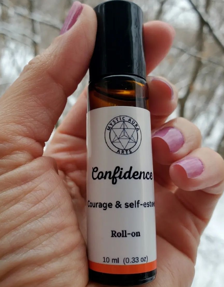 Hand holding a small roll-on bottle labeled 'Confidence' with a tree symbol, outdoors with trees in background.