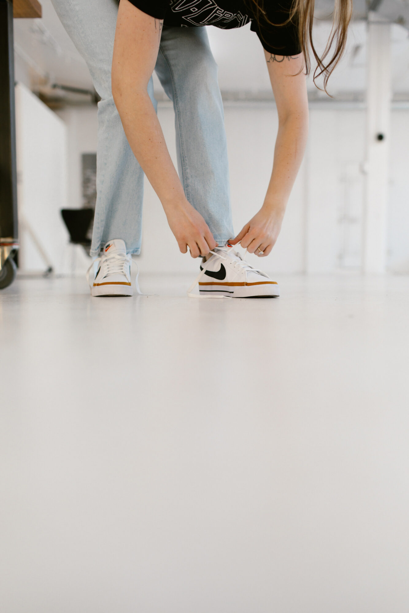 Person bending down to tie shoelaces on white sneakers with black and orange accents, in a bright indoor space.