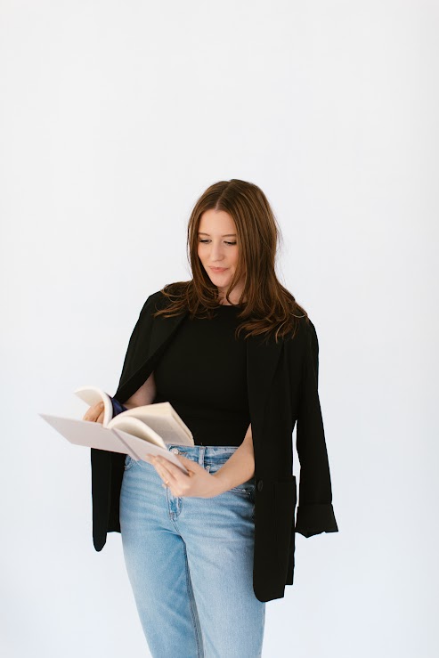 Woman with brown hair reading a book, wearing a black top and light blue jeans, against a white background.