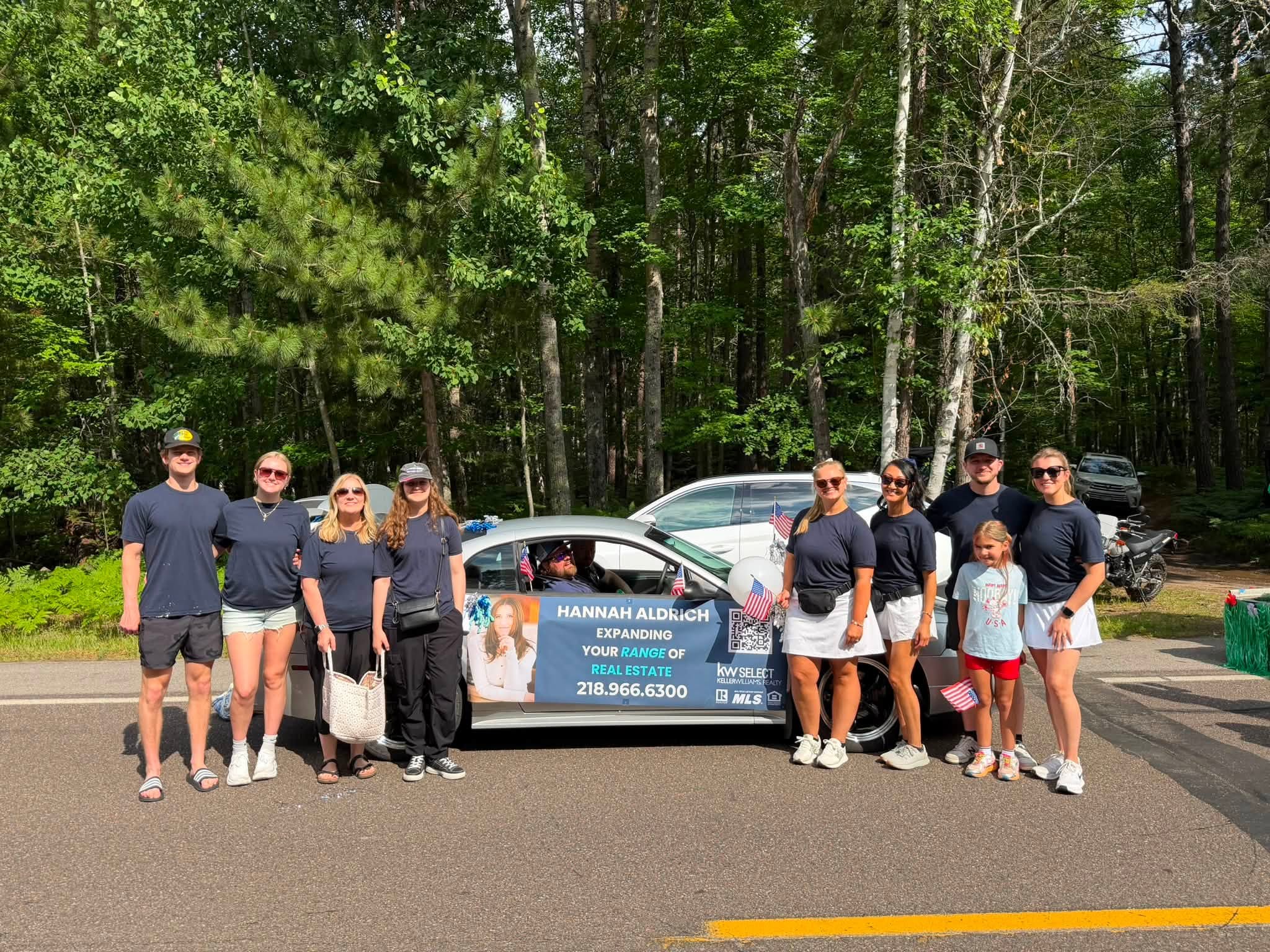 Group of nine people standing outdoors in front of a car with a banner, trees in the background.