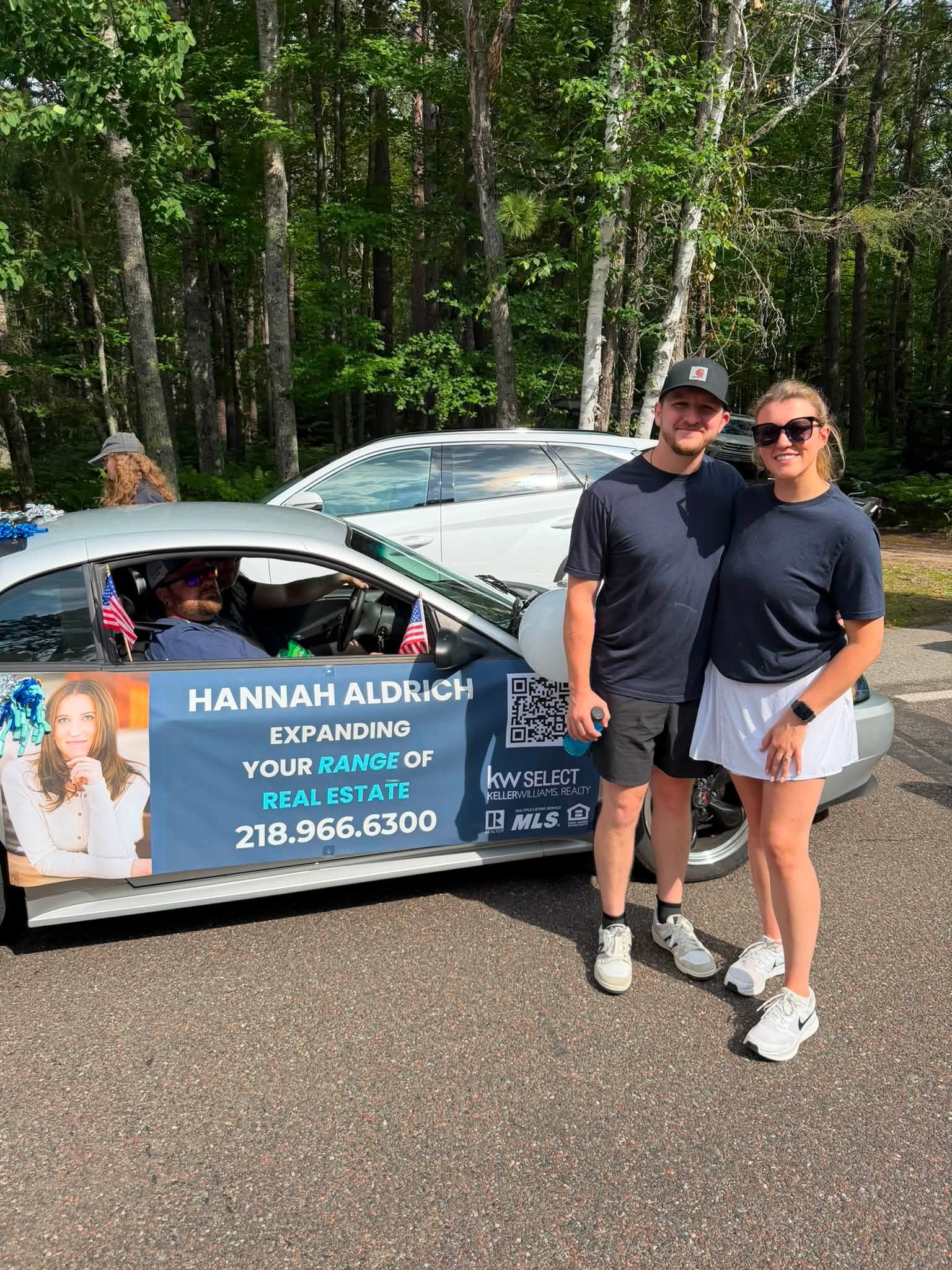 Two people stand next to a car with a real estate sign, in a wooded area, smiling at the camera.