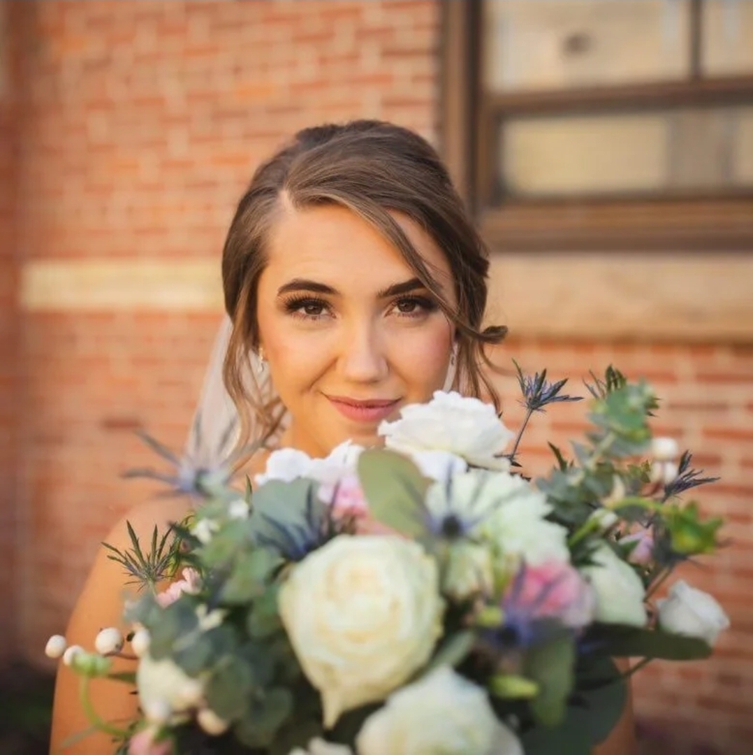 Young woman holding a bouquet of white and purple flowers, smiling, with a brick building in the background.